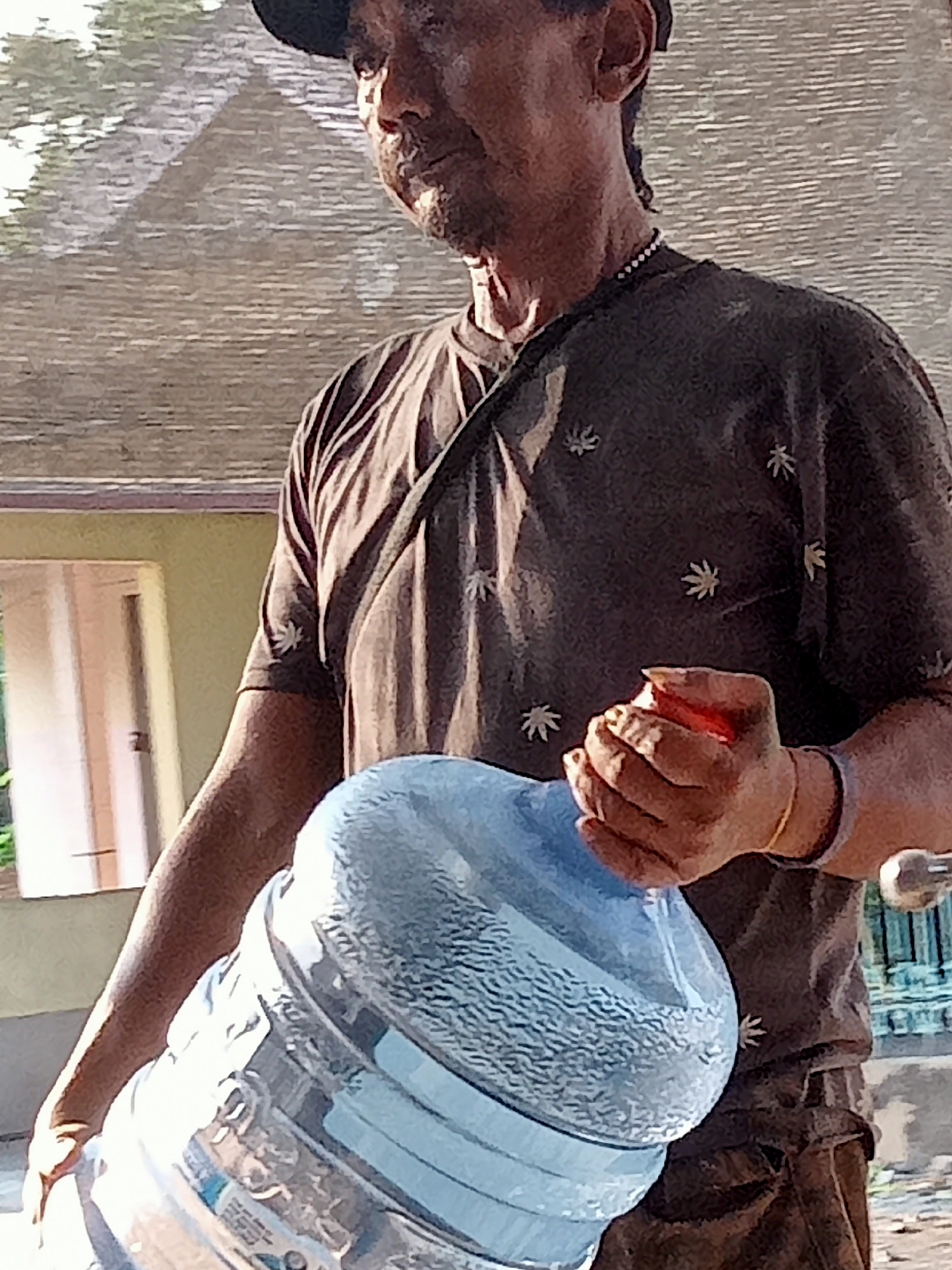 A man wearing a dark t-shirt with a white leaf pattern and a dark baseball cap is holding a large clear plastic water jug. He has a dark complexion, a mustache, and is wearing a beaded necklace. The background shows a building with a beige wall and a light brown roof. The man is in Simo, Indonesia.