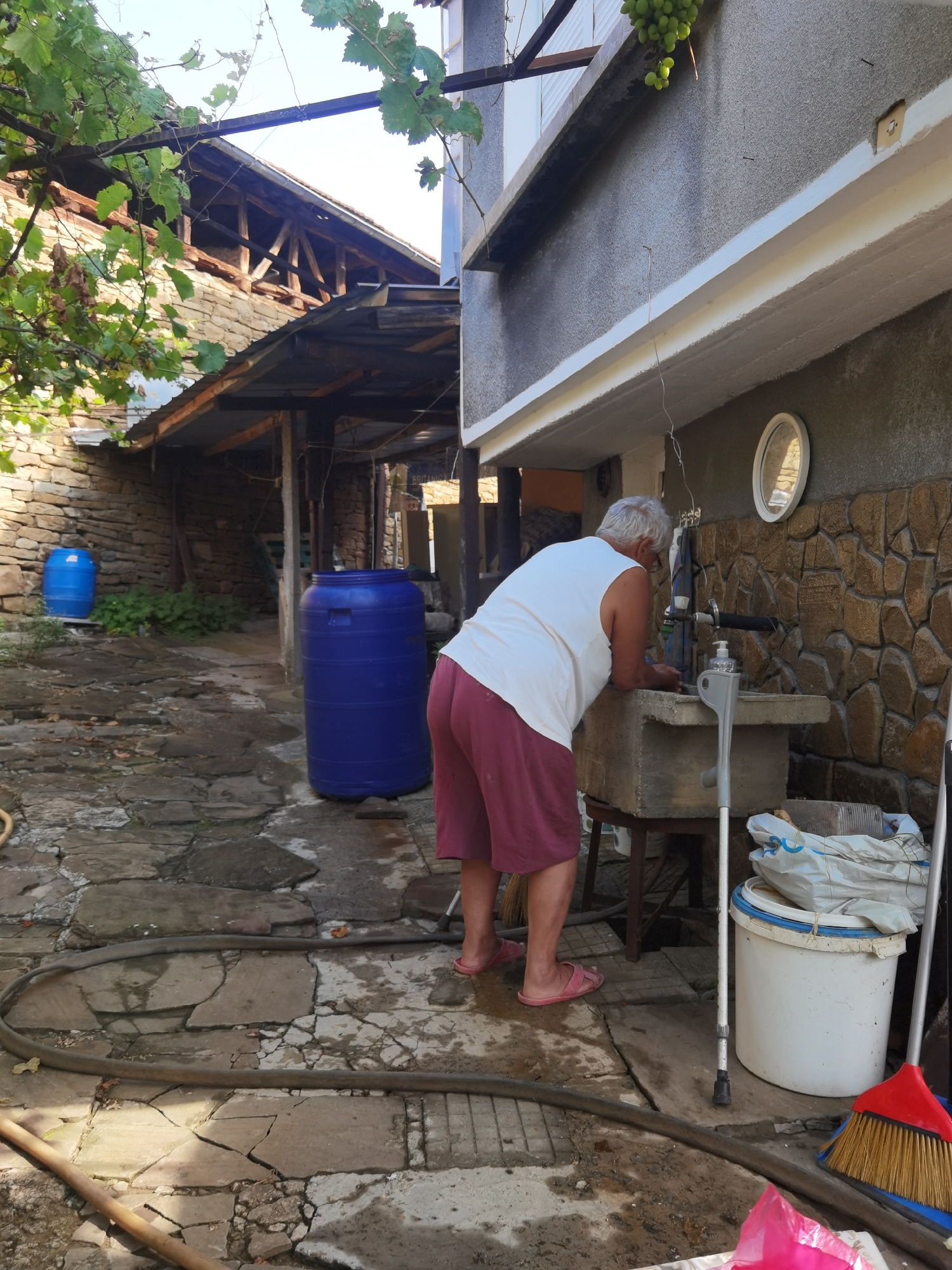 The image depicts an outdoor scene in the village of Lomets, Bulgaria. An elderly man with gray hair, wearing a white sleeveless shirt and maroon shorts, is leaning over a concrete sink, appearing to wash his hands. A grey crutch stands next to the sink, and a white bucket filled with white bags and a blue lid is also present. In the foreground, a dark hose lies on the stone-paved ground. Behind the man, a large blue plastic barrel is visible, and further in the background, another blue barrel is positioned near a stone wall. The architecture suggests a rural setting, with weathered buildings and exposed wooden beams. Green grapevines hang from an overhead structure. The overall impression is of a functional outdoor space, possibly a courtyard or alleyway, in a traditional Bulgarian village.