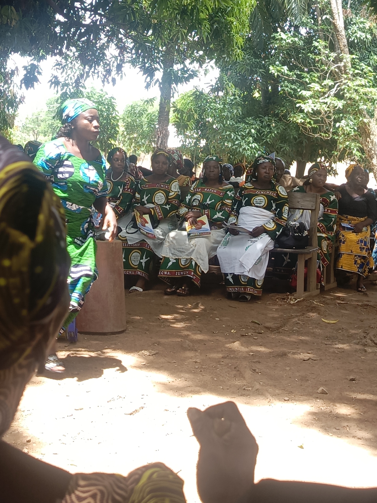 The image shows a gathering of women in a rural setting, likely in or near Ninzo South, Nigeria. Most of the women are wearing matching outfits with green, white, and brown patterns. The text "ECWA ZUMUNTA," "CAR MATAN," and "UMUNTIR ITAUNA" are visible on their dresses. Some women are seated on wooden benches, holding books or papers. One woman stands near a concrete post, addressing the group. The ground appears to be dirt or packed earth. Foliage from trees provides shade. The foreground is somewhat obscured, suggesting the photographer is part of the crowd.