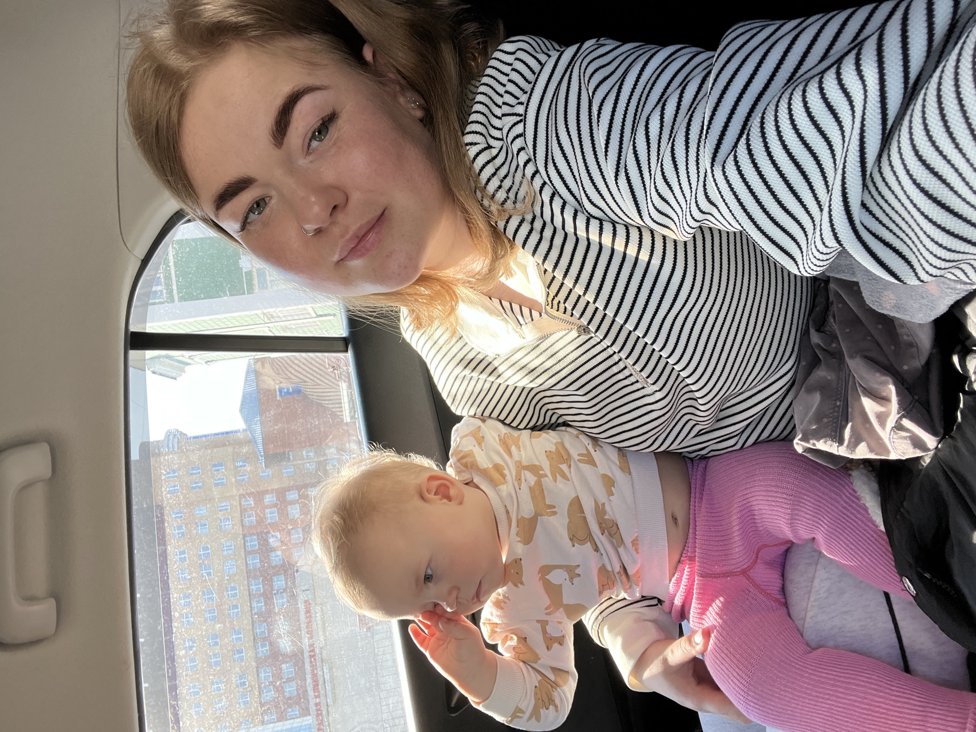 A fair-skinned young woman is holding a baby in her lap inside a vehicle. The woman is wearing a black and white striped hooded jacket. The baby is wearing a white onesie with an animal print and pink leggings. Outside the vehicle window, there is a view of a building in Perm, Russia.