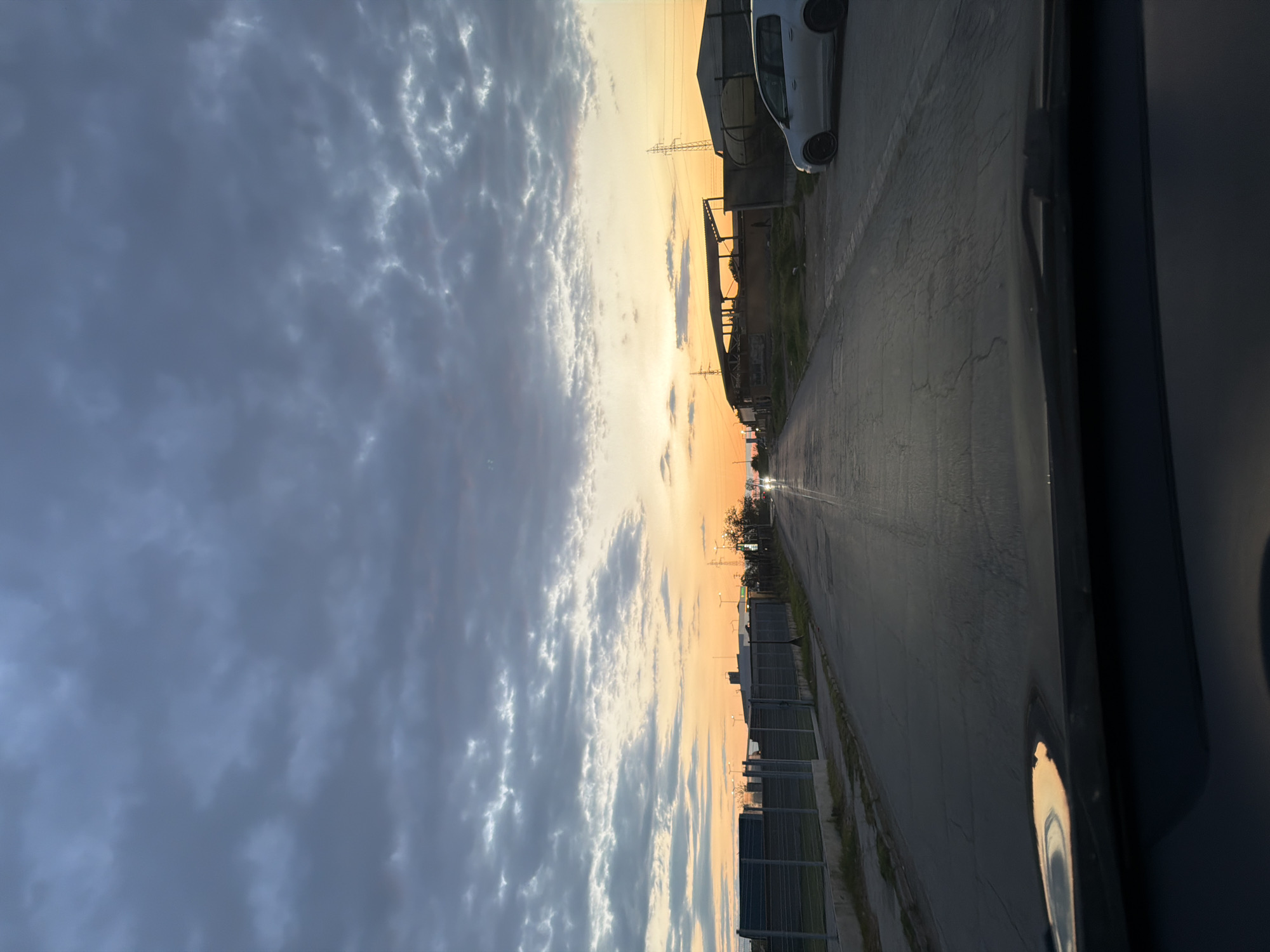 The image, taken from inside a vehicle, shows a street in Parvomay, Bulgaria, during a sunset. The sky is filled with dark clouds tinged with orange from the setting sun. A street runs straight ahead, with buildings on either side, one including a parked white car. Power lines are visible overhead. A light source, potentially a car or street lamp, is seen in the distance on the road. A guardrail flanks the street. The car interior is partially visible in the lower right of the frame.