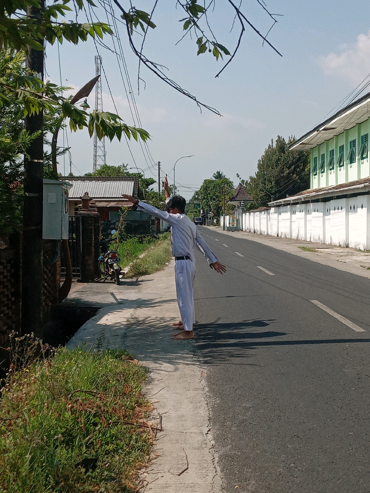 A young person, wearing a white uniform, stands on a concrete sidewalk next to a paved road. Their right arm is extended upwards and to the side, with their hand open, while their left arm is also extended to the side. The person appears to be facing away from the camera. The sidewalk is lined with green vegetation. Across the road, there is a white wall with a green building behind it. In the background, a utility pole with wires is visible, along with trees and houses. A motorcycle is parked on the left side of the image, near a brick fence and an electrical box. The sky is clear and blue. The scene appears to be in a suburban or rural area of Srimulyo, Indonesia, with evidence of infrastructure and residential buildings.