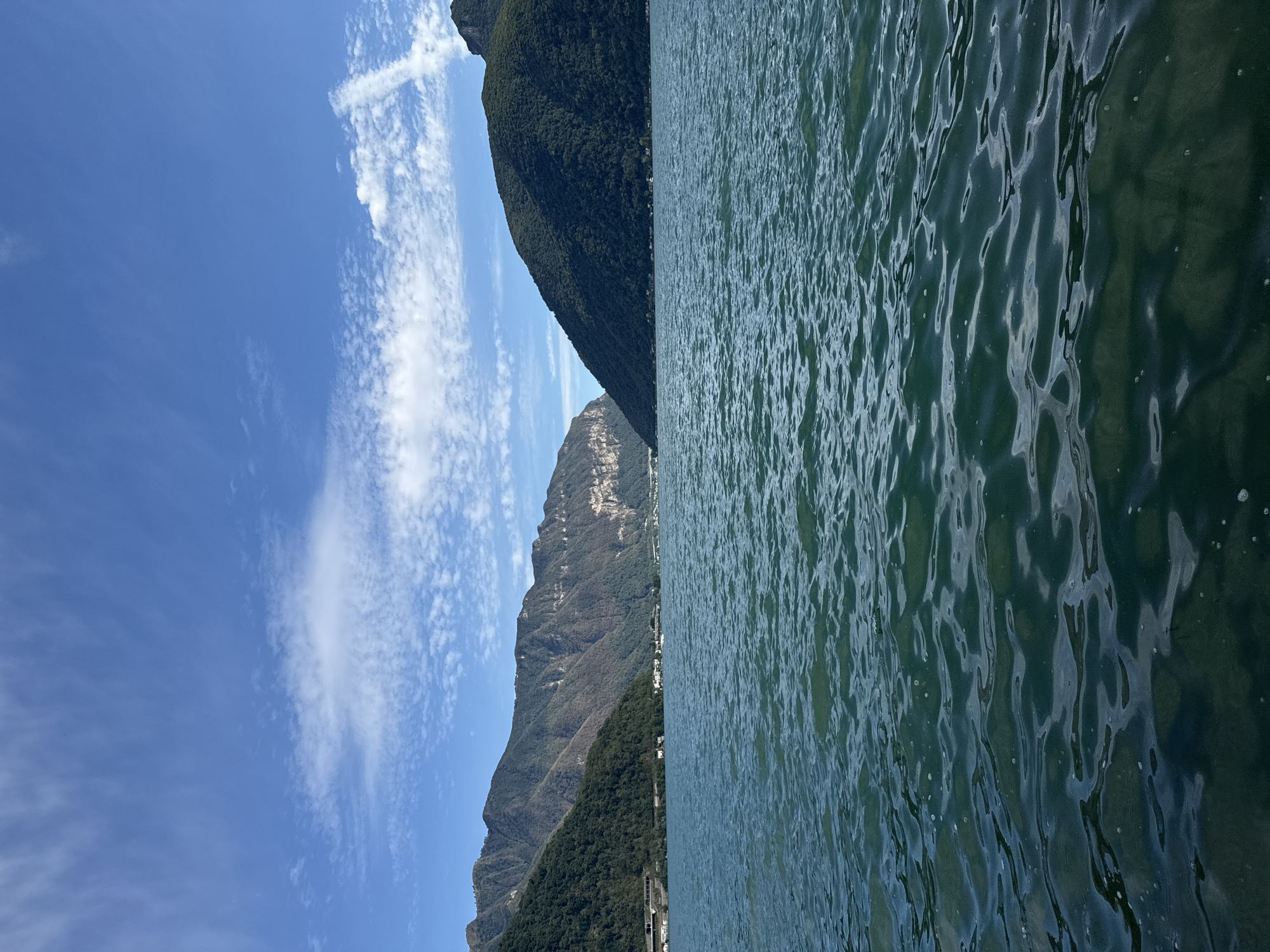 The image displays a serene view of Lake Lugano from the shore in Melide, Switzerland. The foreground is dominated by the deep green, rippling water of the lake, with sunlight creating patterns of light and shadow on its surface. In the middle ground, a cluster of white buildings, indicative of a small town or village, is nestled at the base of lush, tree-covered mountains. The mountains themselves rise steeply, forming the backdrop to the scene. The sky above is a bright, clear blue, punctuated by scattered white clouds, some of which are wispy and elongated, suggesting a gentle breeze. The overall impression is one of natural beauty and tranquility.