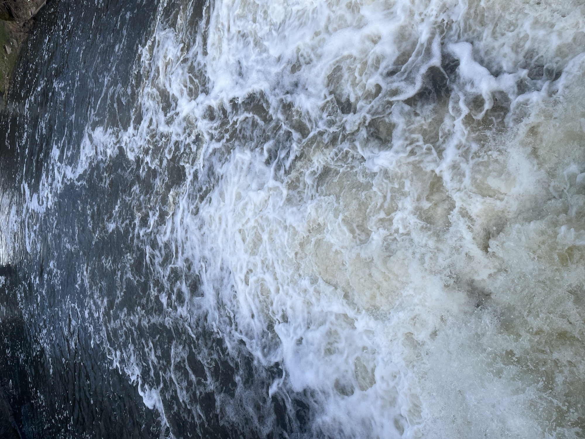 The image shows a turbulent body of water, likely a river or waterfall. On the left, the water appears darker and smoother, transitioning into a frothy, white expanse of churning water on the right. The texture indicates a significant amount of aeration and movement. The location is Klin, Russia. No people are visible in the image.