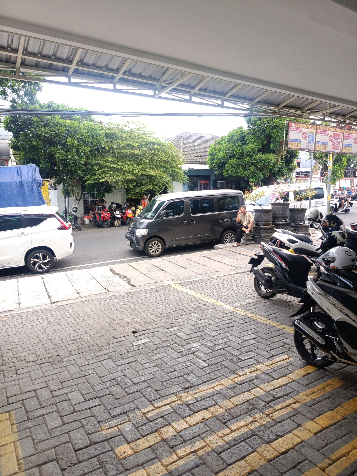 The image depicts a street scene in Kepolorejo, Indonesia, likely in a commercial or residential area with a covered walkway. A dark grey minivan is parked on the road, partially obscuring a white SUV parked to its left. The road is paved with bricks, and yellow lines mark parking spaces. Several motorcycles are parked on the side of the road, some in front of a building with an open storefront where a man is seated on a low wall. Behind the vehicles and buildings, lush green trees are visible. In the background, a white van is also parked, and a person is riding a motorcycle. Advertising banners hang from a structure above the street. The overall impression is of a busy, everyday street in Indonesia.