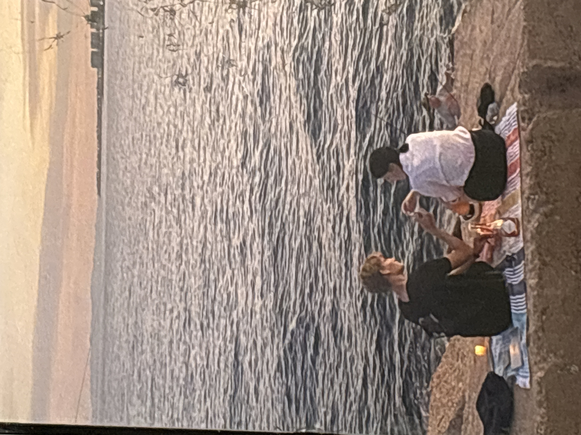 The image depicts a serene sunset scene by the sea. Two individuals are seated on a blanket on a rocky shore, facing the water. A man in a black t-shirt is on the left, and a person in a white shirt and dark pants is on the right. They appear to be engaged in conversation or sharing a moment together. A picnic blanket is spread out, with a few items on it, including what looks like a bottle and a candle. The water is calm, with gentle ripples reflecting the soft light of the setting sun. In the distance, a city skyline is visible along the horizon, suggesting an urban proximity to this natural setting. The overall atmosphere is peaceful and romantic. The location is described as being in Orihuela, Spain, a coastal city known for its beautiful beaches and scenic views.