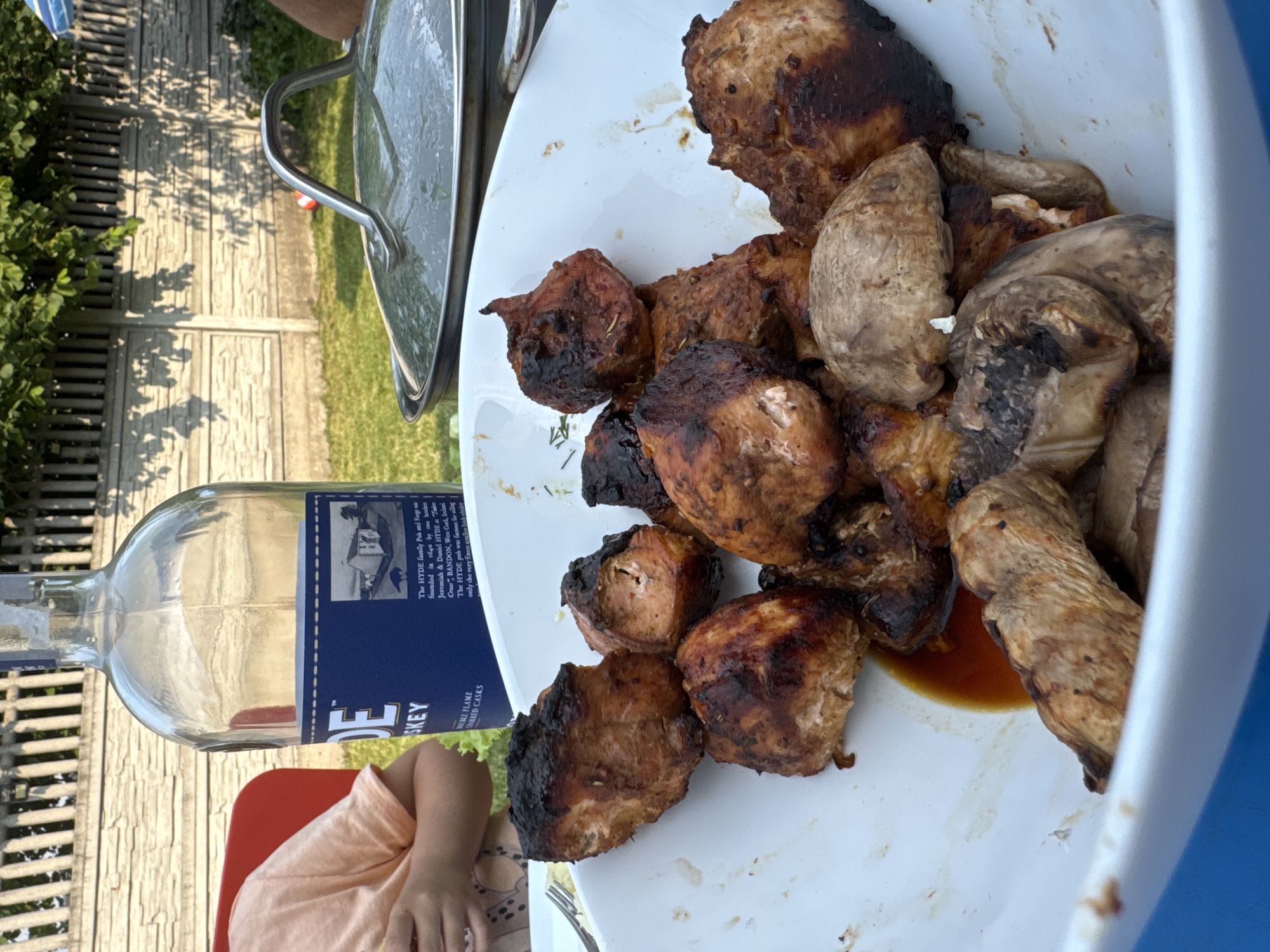 The media file shows a close-up of a white plate filled with grilled chicken and mushrooms. The chicken pieces are varied in size and shape, some are darker and appear well-charred, while others are lighter brown. Herbs, possibly rosemary, are visible on some of the chicken pieces. The mushrooms are large and appear to be whole or halved. Some brown sauce is pooled on the plate.
To the left of the plate, a clear glass bottle with a blue label is partially visible. Text on the label is legible in the cropped image, indicating it's a whiskey bottle. The text reads: "DE SKEY DOUBLE FLAME OAK AGED CASKS" and describes "The HYDE family Pub and Forge" founded in 1640 by brothers Jeremiah and Daniel Hyde in Bandon, West Cork, Ireland, famous for "only the very finest quality Irish whiskey".
In the background, out of focus, is a large pot with a metal handle, suggesting it might be a cooking pot or a serving dish with a lid. A portion of a person's arm and hand is visible in the lower left, wearing a light-colored, possibly peach-colored, short-sleeved shirt. The setting appears to be outdoors, with a wooden fence and greenery visible in the background. Sunlight casts shadows, indicating it is daytime. The overall scene suggests a casual outdoor meal, possibly a barbecue or picnic.