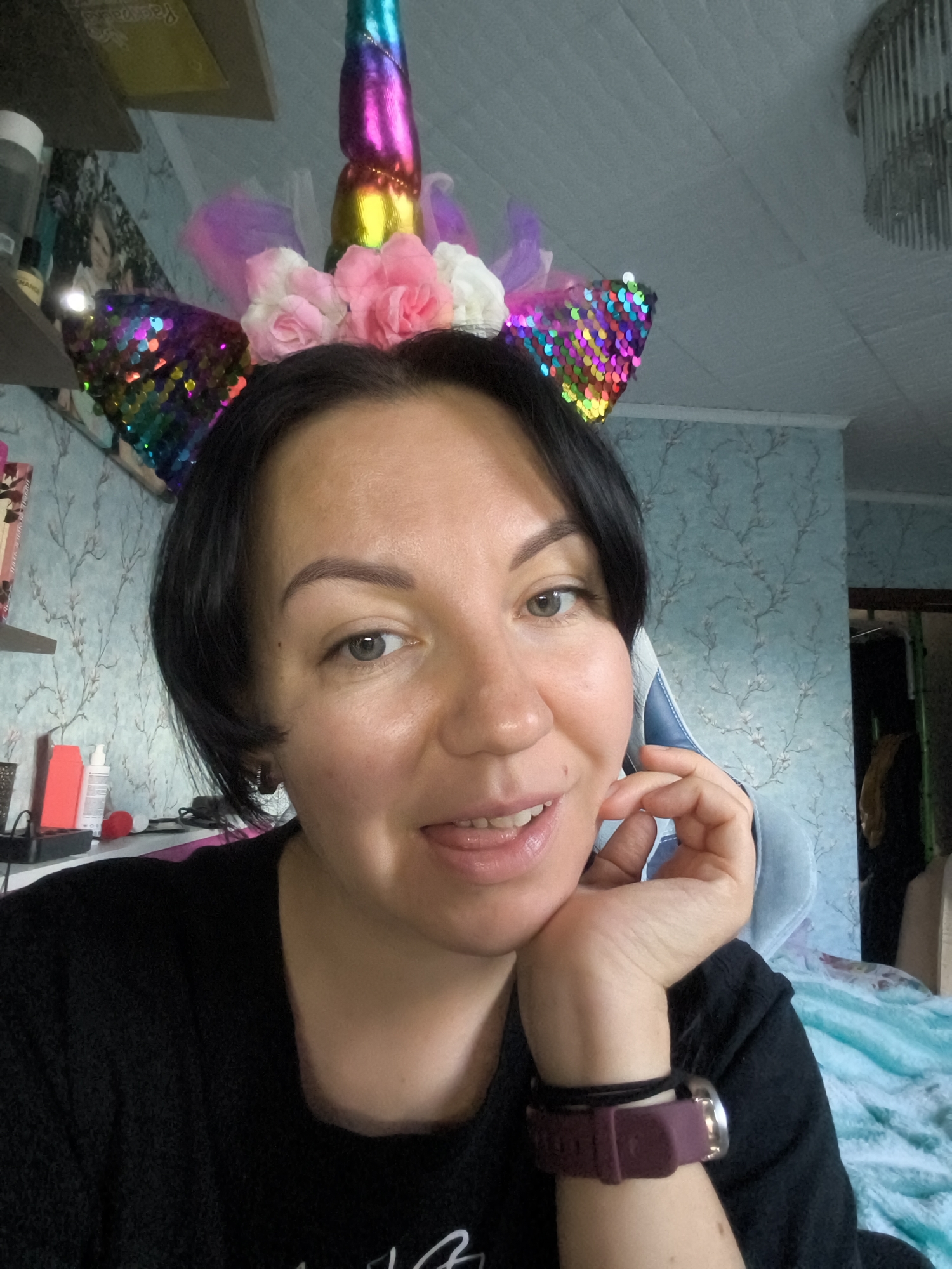 A medium shot shows a woman with dark hair wearing a black shirt. She is looking at the camera with her chin resting on her hand. She has on a unicorn horn headband with colorful sequins and flowers. In the background, there is blue patterned wallpaper and a chandelier. The scene appears to be indoors, possibly in a home.