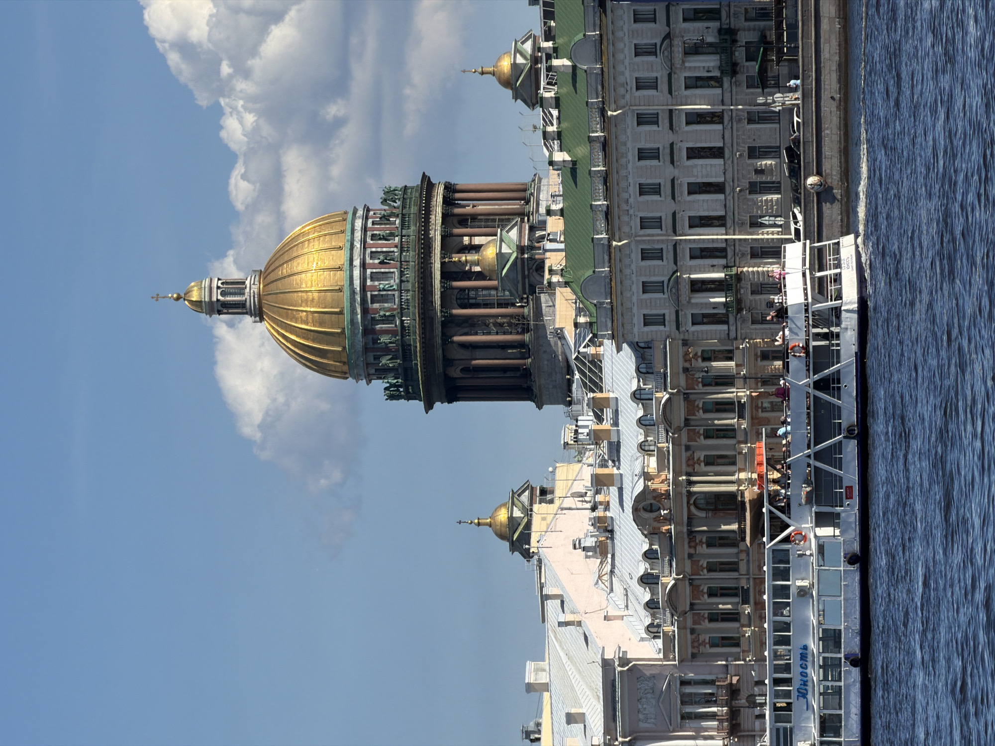 This panoramic image depicts a vibrant daytime scene in Saint Petersburg, Russia, showcasing the iconic architecture along a wide river or canal. The most prominent feature is the magnificent golden dome of what appears to be Saint Isaac's Cathedral, positioned on the left side of the frame. The cathedral dome is highly ornate, featuring fluted gold panels, surrounded by a colonnade with classical columns and intricate sculptural details and smaller domes at its base.
To the right of the cathedral, a series of grand, multi-story buildings with classical facades, numerous windows, and green-colored roofs line the embankment. Some of these buildings also feature smaller golden domes, echoing the cathedral's grandeur.
In the foreground, a white tour boat is visible on the water, docked or moving alongside the embankment. Several people are seen on the boat's open upper deck, likely enjoying a sightseeing cruise. A life preserver is mounted on the boat's railing. The water itself is a deep blue, showing ripples on its surface. The name "ЮНОСТЬ" (Yunost, meaning "Youth") is clearly visible in blue lettering on the side of the boat.
The sky above is bright blue with large, fluffy white clouds, indicating clear and pleasant weather. The overall impression is one of a sunny day, ideal for exploring the city's historic waterways and architectural marvels.