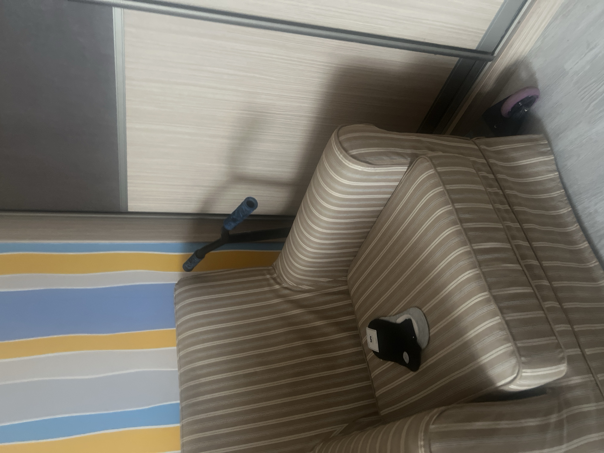 The image depicts an indoor scene, likely a living space. In the foreground, a striped armchair upholstered in beige and white fabric occupies the left side of the frame. On the seat of the armchair, a pair of black and grey socks are placed, one on top of the other. A scooter with blue handlebars is positioned behind the armchair, leaning against the wall. The wall behind the armchair features a vertical striped wallpaper in alternating colors of yellow, blue, and white. To the right of the armchair, a large wardrobe with light-colored wooden doors is visible. A single purple wheel, possibly from a roller skate or similar item, is on the floor near the base of the wardrobe and the armchair. The floor appears to be a light-colored laminate or wood. The image does not provide any specific visual cues to pinpoint the location within Surgut, Russia.
