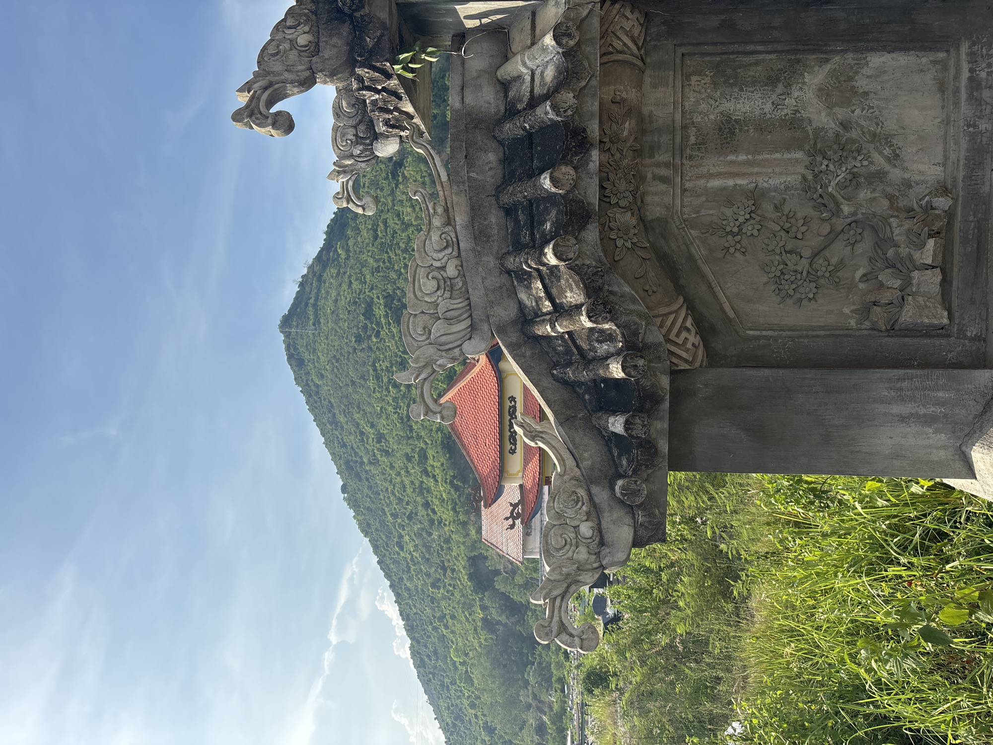 The image shows a stone carving in the foreground, with a lush green mountain and a temple in the background. The stone carving features intricate details, including what appear to be dragons or mythical creatures on the roofline and a floral relief on the side. The mountain behind is covered in dense vegetation, and a clear blue sky with scattered clouds stretches above. A temple with a red-tiled roof is visible nestled on the slope of the mountain. The scene suggests a tranquil, possibly religious or historical site, likely located in a natural setting. The location is specified as Tay Nha Trang Ward, Vietnam.
