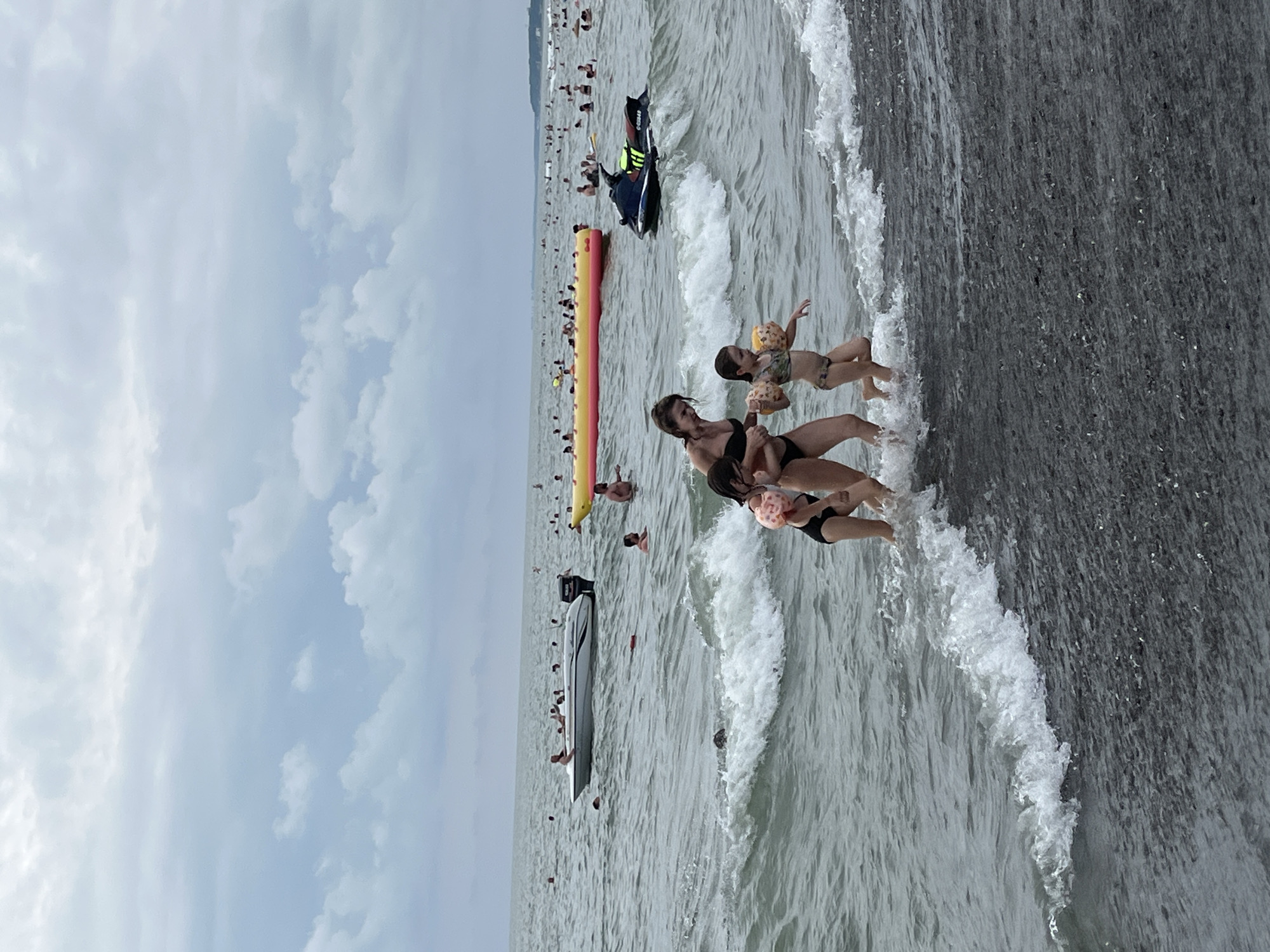 This media file depicts a lively scene at Ureki, Georgia, a location known for its magnetic black sand beaches. The image is oriented vertically and captures a bustling beach environment.
The primary subjects in the foreground are two people, a woman and a child, standing in shallow water with gentle waves lapping at their feet. The woman is wearing a black bikini, and the child, on her right, is wearing a patterned bikini and arm floaties. They appear to be enjoying a day at the beach, with the child holding a pink inflatable object.
In the background, the sea is populated with numerous individuals swimming and enjoying various water activities. Several objects are visible in the water, including a long, yellow and red inflatable tube, commonly known as a banana boat, and a sleek, white speed boat moored in the distance. A jet ski is also visible, creating a foamy wake as it moves across the water. The water itself is a muted grey-green, with visible waves and ripples.
The weather appears to be overcast, with a light grey sky filled with scattered clouds dominating the upper portion of the frame. This suggests it might be a cloudy day, or possibly late afternoon as the light seems diffused. The overall atmosphere is one of leisure and recreation, with many people engaging in typical beach activities. The presence of the speed boat, jet ski, and inflatable tube indicates a vibrant tourist destination offering various forms of water entertainment. No visible text is present in the image.