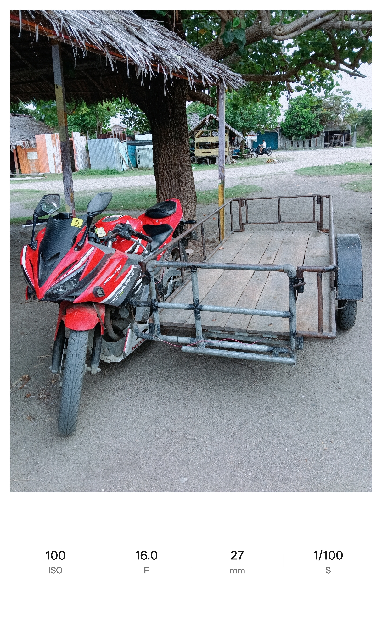 A red motorcycle with a sidecar is parked on a sandy surface under a thatched roof structure. The motorcycle has a black windscreen, and the sidecar is made of metal and wood. In the background, there are several huts and trees. A motorcycle is seen in the distance on the road. The image was likely taken in daylight.