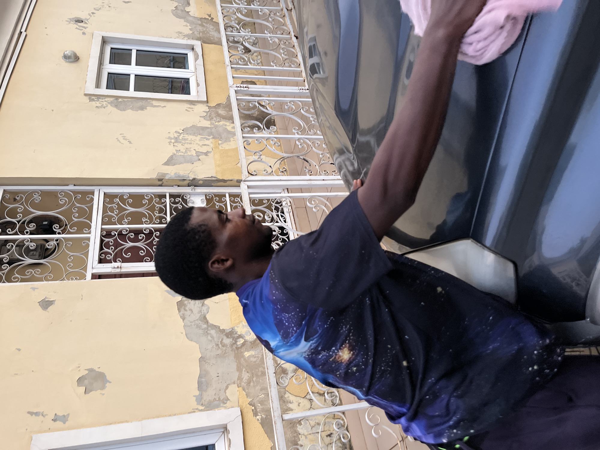 In Abuja, Nigeria, a man with dark skin and short black hair is seen from the chest up, facing to the right and leaning over to polish a dark-colored car with a pink cloth. He is wearing a dark blue t-shirt adorned with a celestial pattern featuring stars and nebulae. The background of the image consists of a weathered, pale yellow building with peeling paint. Two windows are visible: one on the left with a white frame, and another on the right, also with a white frame, showing a crossbar design. A white metal railing with decorative scrollwork runs horizontally across the middle ground, separating the building from the car.