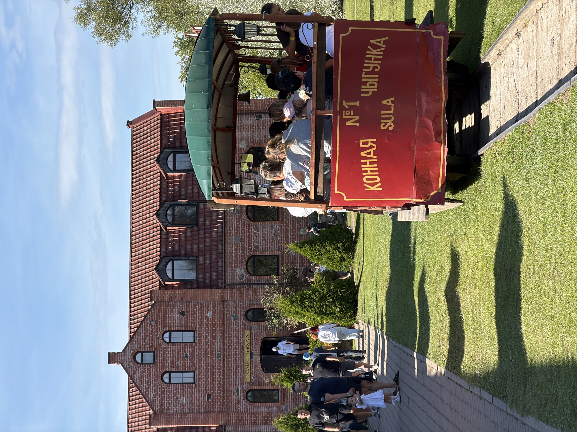 In the city of Літвенскі сельскі Савет, Belarus, a red train car, part of a tourist attraction, is captured in a detailed shot. The train car, featuring a green roof and ornate lanterns, is filled with people enjoying a scenic ride. The car's side displays Cyrillic text, "КОННАЯ N°1 ЧЫГУНКА SULA," indicating it's a horse-drawn railway. The background reveals a charming brick building with arched windows and a red tiled roof, characteristic of the region's architecture. A paved walkway leads to the building, lined with lush green grass and conical evergreen trees. The scene is bathed in bright sunlight, casting long shadows across the lawn, indicating a clear day. The overall impression is one of a pleasant outdoor excursion in a picturesque Belarusian setting.