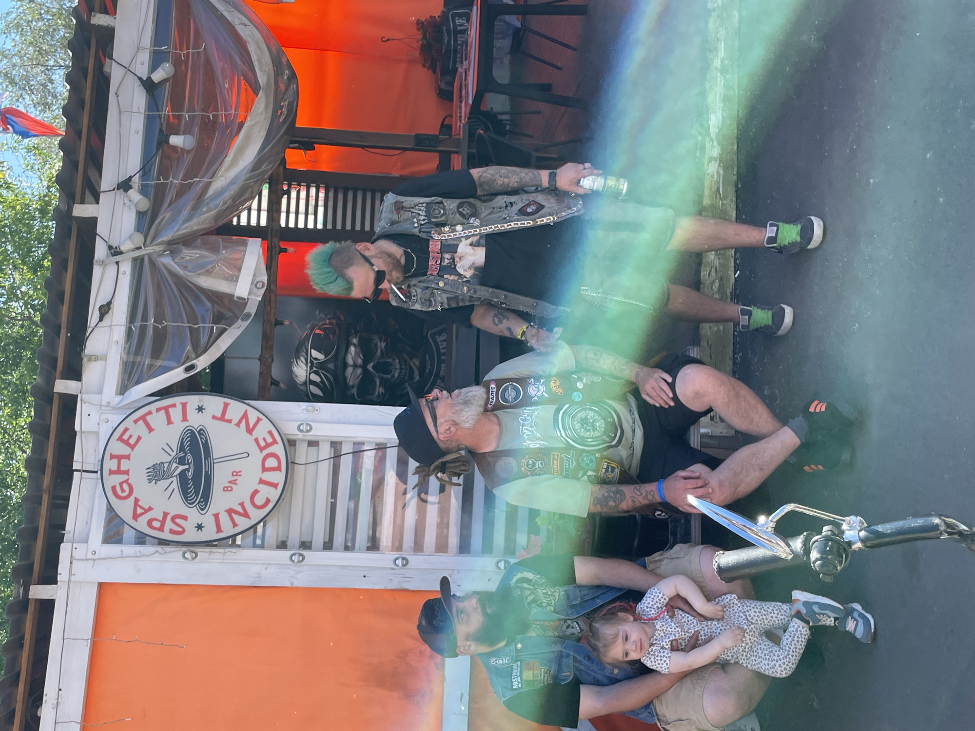 The image captures a group of people gathered outside a building with an orange facade and a sign that reads "SPAGHETTI BAR INCIDENT." The scene appears to be outdoors, possibly a bar or a casual gathering spot. The lighting suggests it is daytime, with some sunlight creating lens flares across the image.
On the left, a man with a beard and a black baseball cap is sitting, holding a young child on his lap. The child is wearing a patterned onesie and has red hair ties. The man is wearing a denim vest over a t-shirt.
Beside him, another man with a beard and dreadlocks is seated, wearing a black hat and glasses. He has a patch on his vest that reads "KRAZIES."
To the right of these two men stands a person with bright green mohawk hair, sunglasses, and a cigarette in their mouth. They are wearing a denim vest decorated with various patches and chains. They are holding a can in their right hand.
The background shows elements of the building's exterior, including plastic sheeting, string lights, and what appears to be tables and chairs further back, suggesting an outdoor seating area. The overall atmosphere seems informal and relaxed.
The location is identified as Saint Petersburg, Russia.