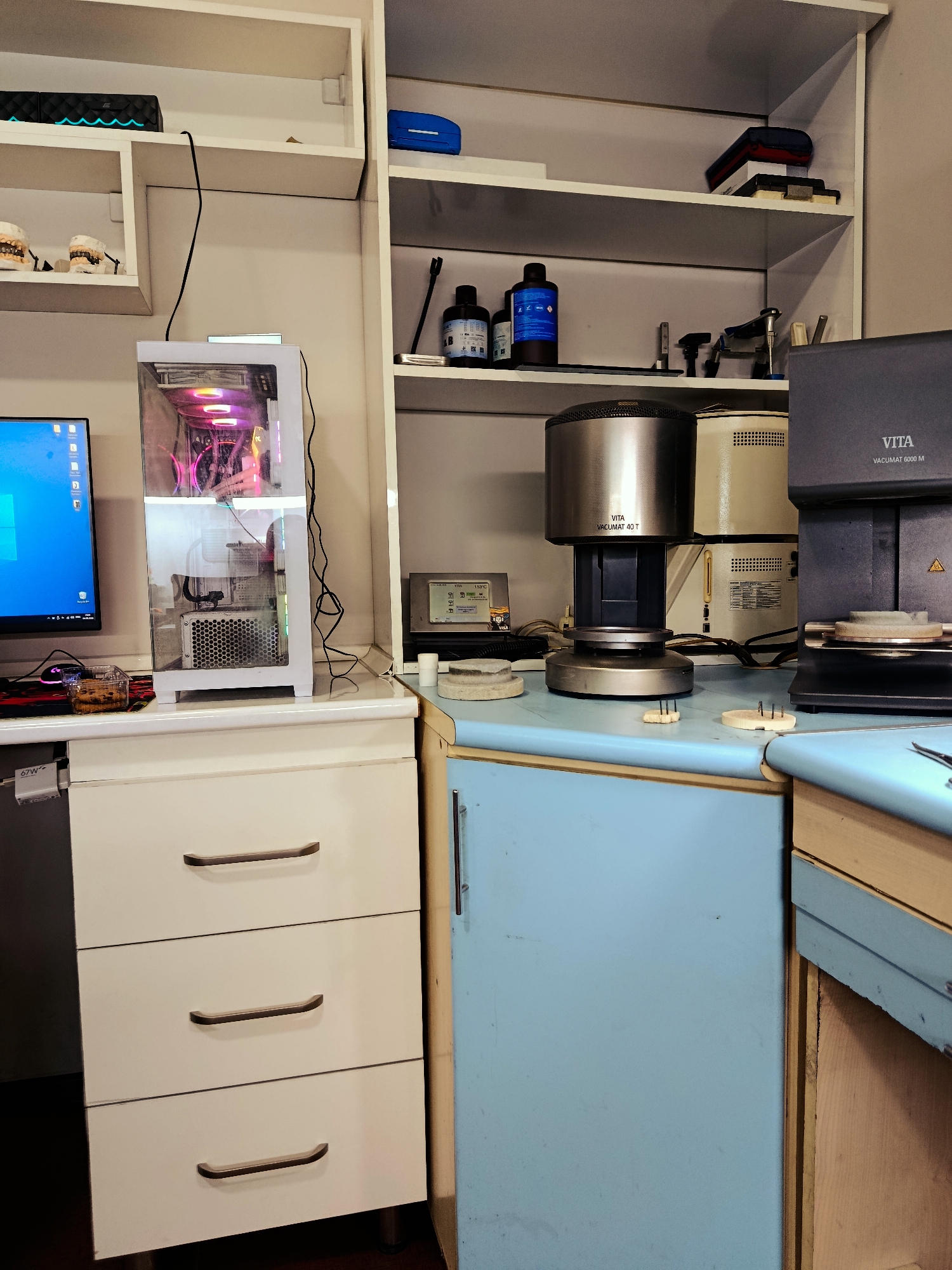 The image depicts a laboratory or workshop environment, possibly a dental laboratory, located in Ganja City, Azerbaijan. On the left, a white computer tower with RGB lighting sits on a desk next to a computer monitor displaying the Windows operating system. Below the monitor is a mousepad with a mouse and a small transparent container with what appears to be liquid and a small object inside. To the left of the computer, on a shelf, are two dental models, one showing an upper jaw and the other a lower jaw, with what look like porcelain crowns or veneers.
The central and right portions of the image are dominated by various laboratory equipment. A prominent stainless steel furnace, labeled "VITA VACUMAT 40 T", is situated on a countertop. Behind it are two beige units, possibly other dental furnaces or related equipment. To the right of these is a dark gray unit labeled "VITA VACUMAT 6000 M". Various ceramic or impression materials are visible on the countertop in front of the furnaces.
Above the countertop, shelves are visible. The top shelf on the left holds a black electronic device with blue lighting and a blue office supply item. The shelves on the right contain several bottles of what appear to be liquids, likely chemicals or dental materials, along with other lab instruments. A blue cabinet is positioned below the countertop in the center of the image. The overall setting appears to be a functional workspace with equipment for dental prosthetics or ceramic work.