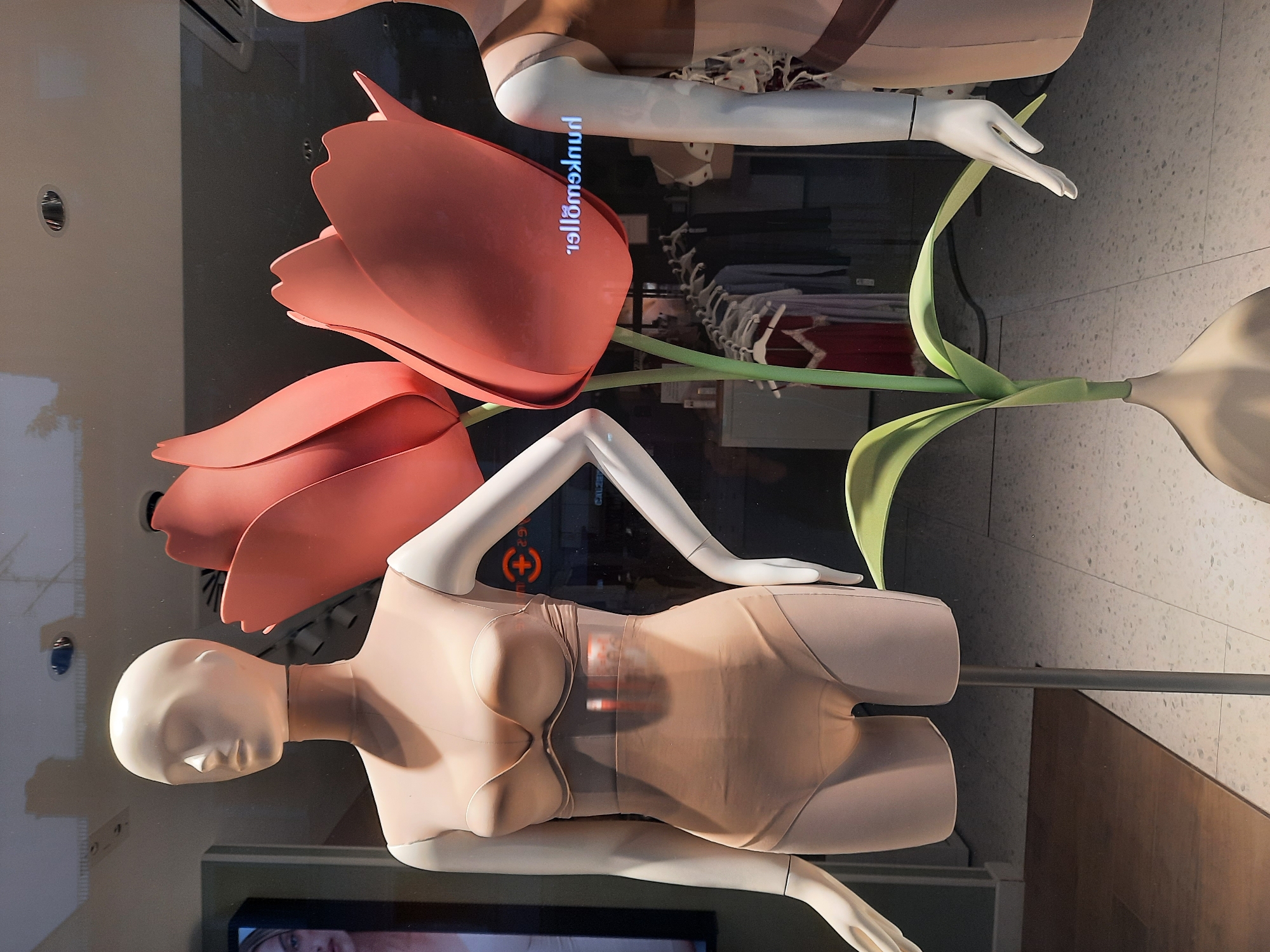 A store window display in Rosenheim, Germany features two mannequins. The mannequin on the left is wearing a nude-colored bodysuit with a built-in bra. The mannequin on the right is partially visible and appears to be wearing a similar garment. In the foreground, a large, stylized pink tulip sculpture stands between the mannequins. The background of the display shows racks of clothing and a reflection of a sign that reads "hellomakeup" in reverse.
