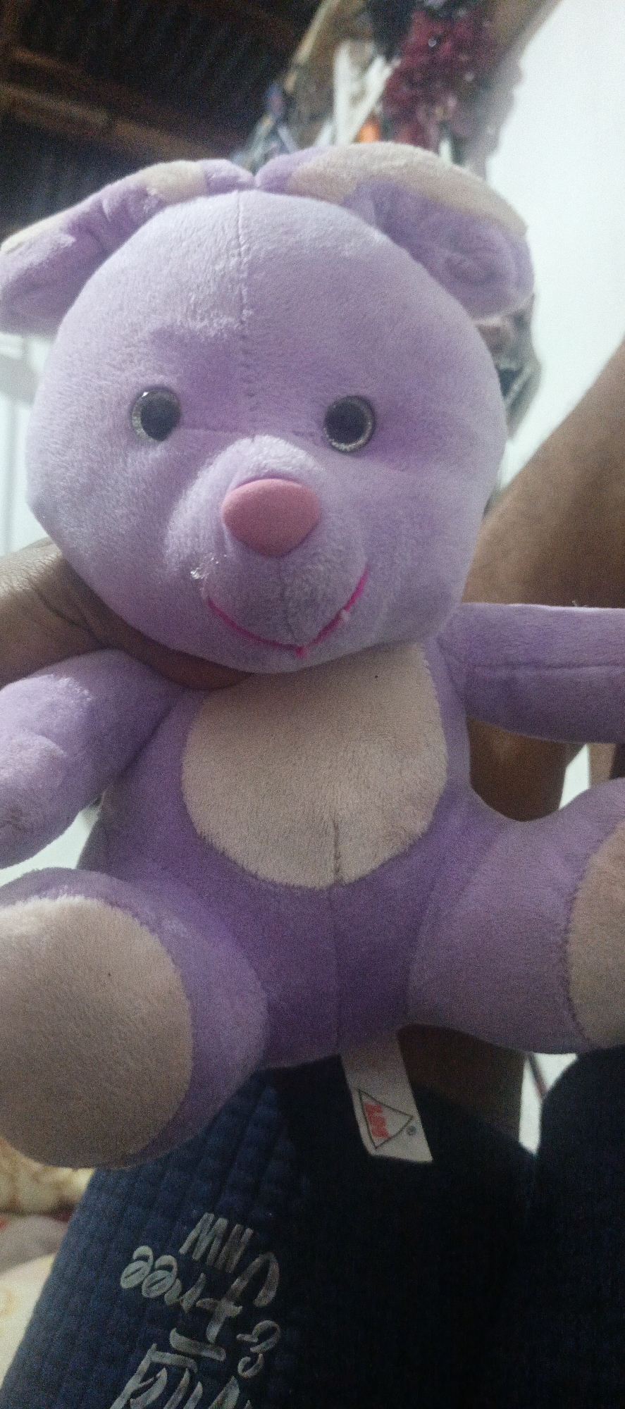 A close-up, slightly blurry, eye-level shot shows a lavender plush toy, possibly a bear or rabbit, being held by unseen hands. The toy has a light pink nose, embroidered pink stitching for a mouth, and shiny black plastic eyes with silver rims. A small wet spot is visible on its snout. The plush toy has lighter colored ear insides and a lighter colored oval on its chest. It is positioned against a dark, out-of-focus background that appears to be indoors. Below the toy, a dark blue fabric, possibly clothing, is visible with some white text that is difficult to decipher due to the blur. A small white tag with a red triangular logo is attached to this fabric. The lighting is dim. The image does not provide any information that allows for localization within Cinta Dame, Indonesia or any specific environment.