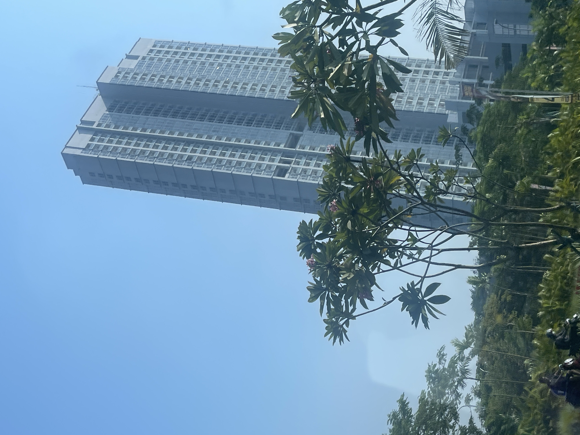 In a bright, clear sky over East Jakarta, Indonesia, a tall, modern skyscraper dominates the background. The building features a repeating pattern of glass windows and light-colored panels, with a stepped architectural design near its summit. In the foreground, lush green trees and foliage partially obscure the view, with some trees bearing light pink flowers. The leaves are large and ovate. Two people on motorcycles are visible on a road in the lower left corner of the image, passing through a well-vegetated area that suggests a park or a landscaped urban environment. The overall scene depicts a blend of urban development and tropical greenery.