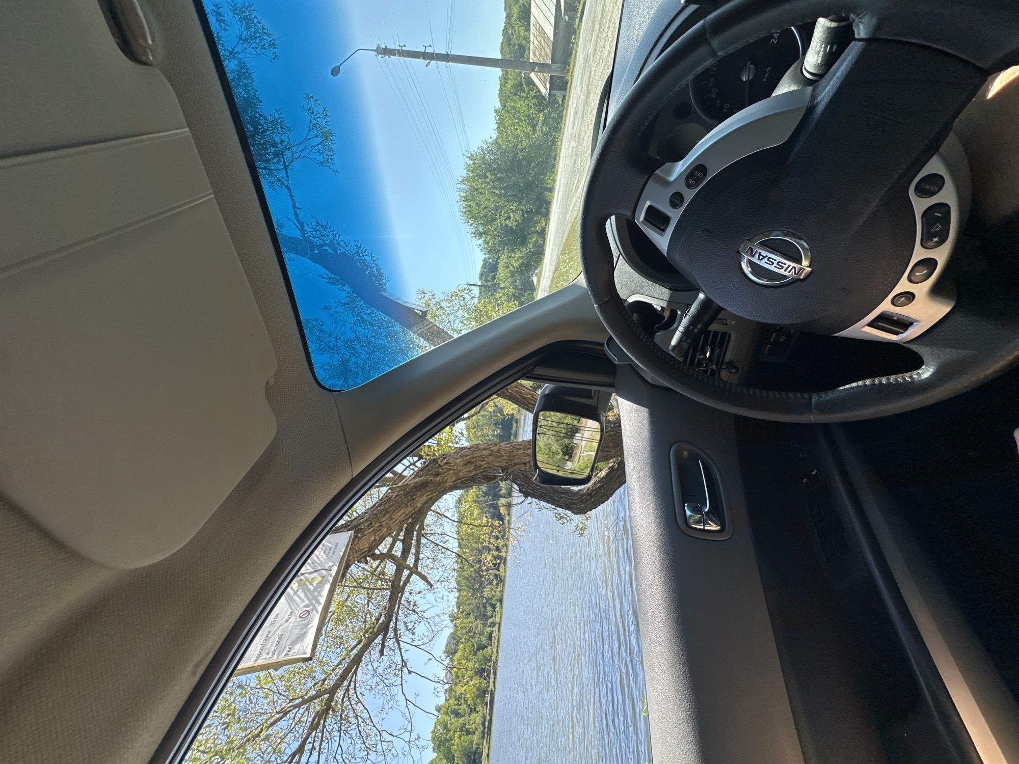 The image, taken from the interior of a Nissan vehicle, shows a view through the open driver's side window and the windshield. To the left, a large lake is visible, bordered by lush green trees. A mature tree with a distinctive curved trunk grows near the water's edge, partially obscuring a road sign. A side-view mirror reflects a paved road and more trees. The steering wheel and dashboard of the car are in the foreground, with the Nissan logo prominently displayed on the steering wheel. The sky is a clear, bright blue. The scene appears to be in or near Makiv, Ukraine, given the context. The environment suggests a rural or semi-rural setting, possibly a roadside park or scenic overlook by a body of water. There are no people visible in the image.