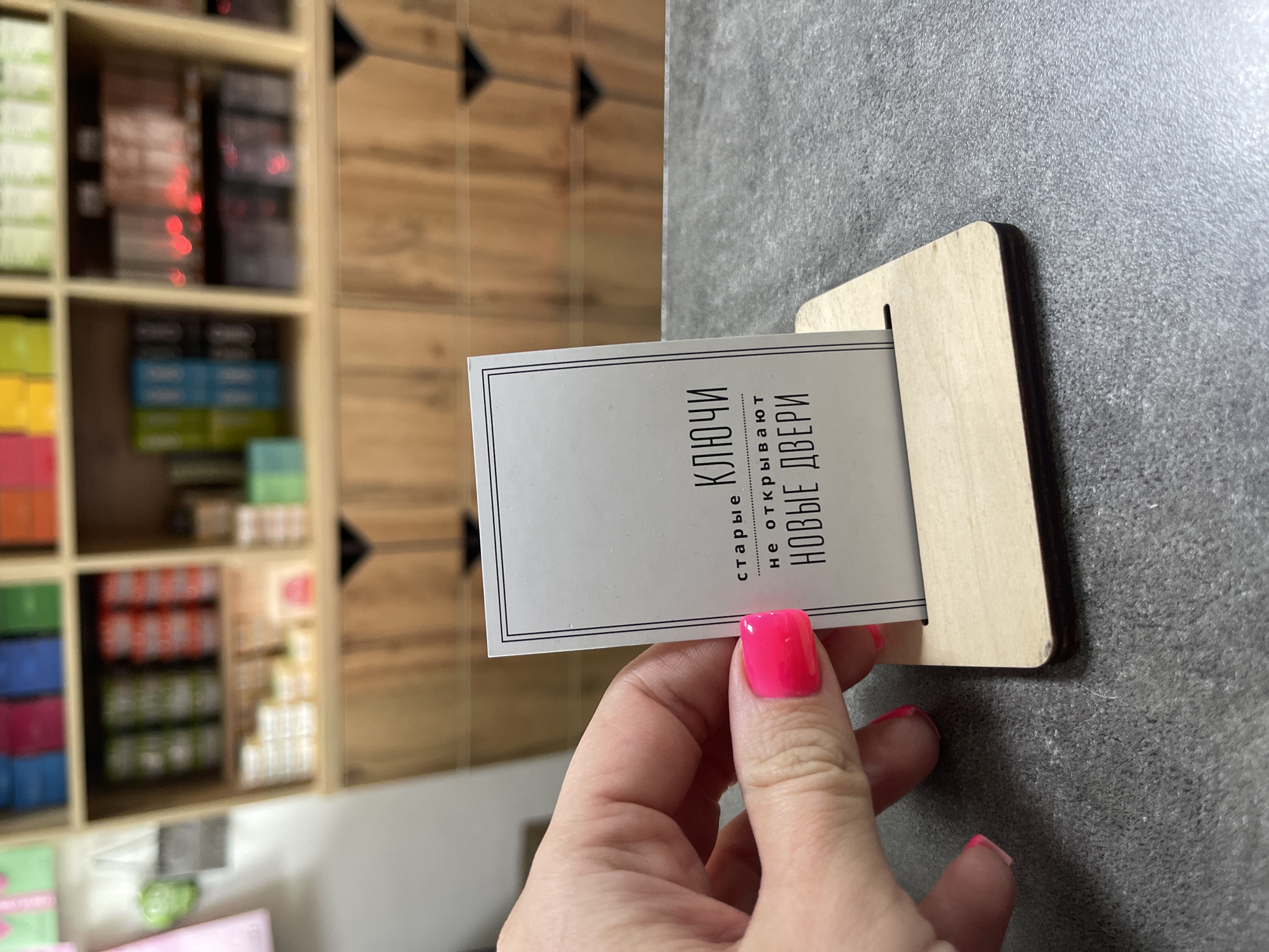 A close-up, eye-level, indoor shot shows a person's hand with bright pink fingernails holding a small, white card with black text, standing upright in a light-colored wooden card holder. The card features the Russian text "старые ключи не открывают новые двери," which translates to "old keys do not open new doors." The background is blurred but appears to be a retail or storage space with shelves filled with various items. The setting does not provide specific details to pinpoint a location in Blagoveshchensk, Russia. The lighting is bright and even, and the focus is sharp on the card and the hand.