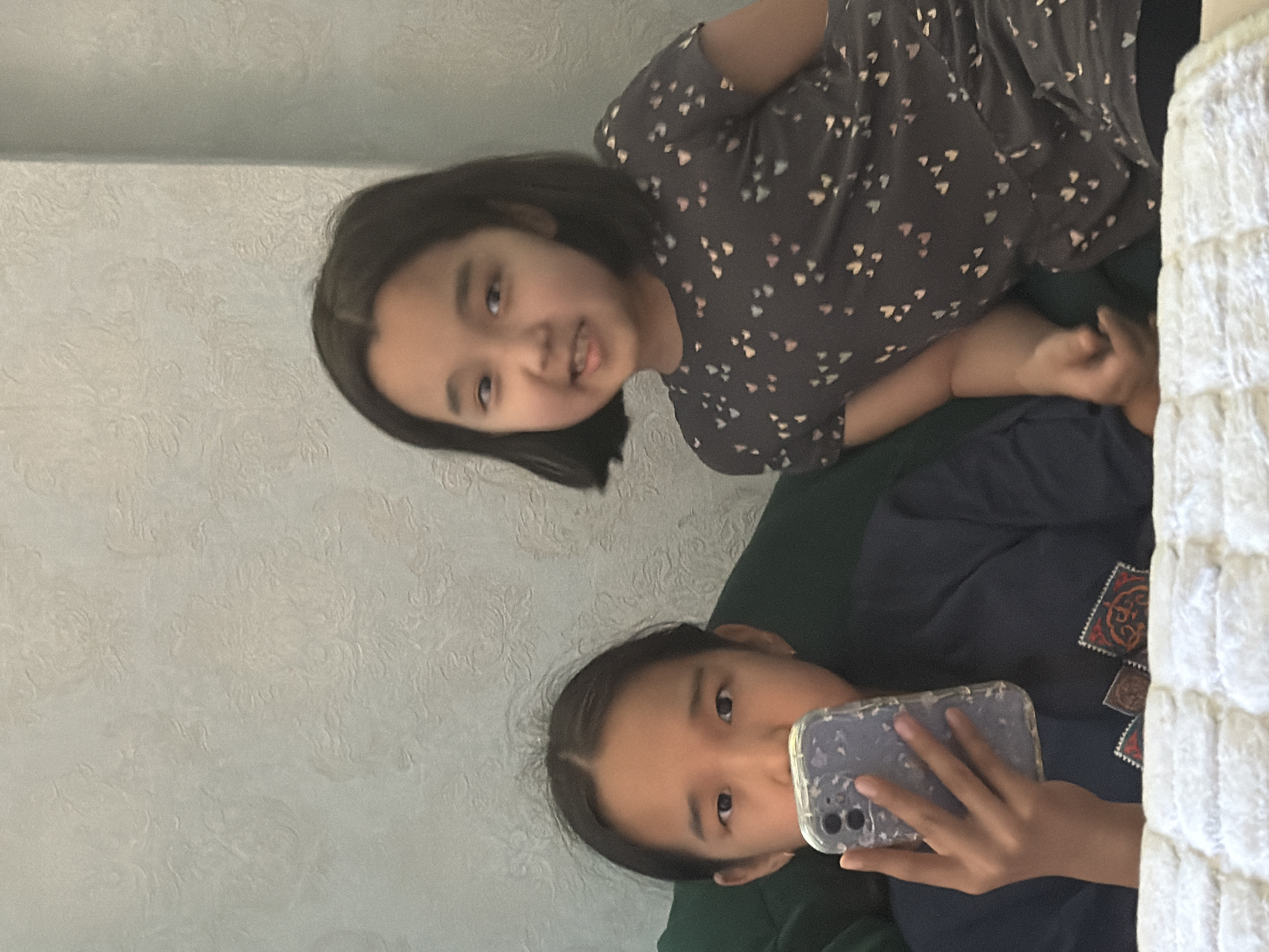 Two young girls are in an indoor setting, likely a bedroom. The girl on the left is closer to the camera, holding a smartphone in her left hand and looking directly at it. She has dark hair pulled back and is wearing a dark blue shirt with a patterned design on the lower front. The girl on the right is slightly behind and to the right of the first girl, looking towards the camera with a slight smile. She has short dark hair and is wearing a dark grey t-shirt with a pattern of small colorful hearts. They are positioned in front of a textured, light-colored wall. The foreground shows a white, textured surface, possibly a bedspread or blanket.