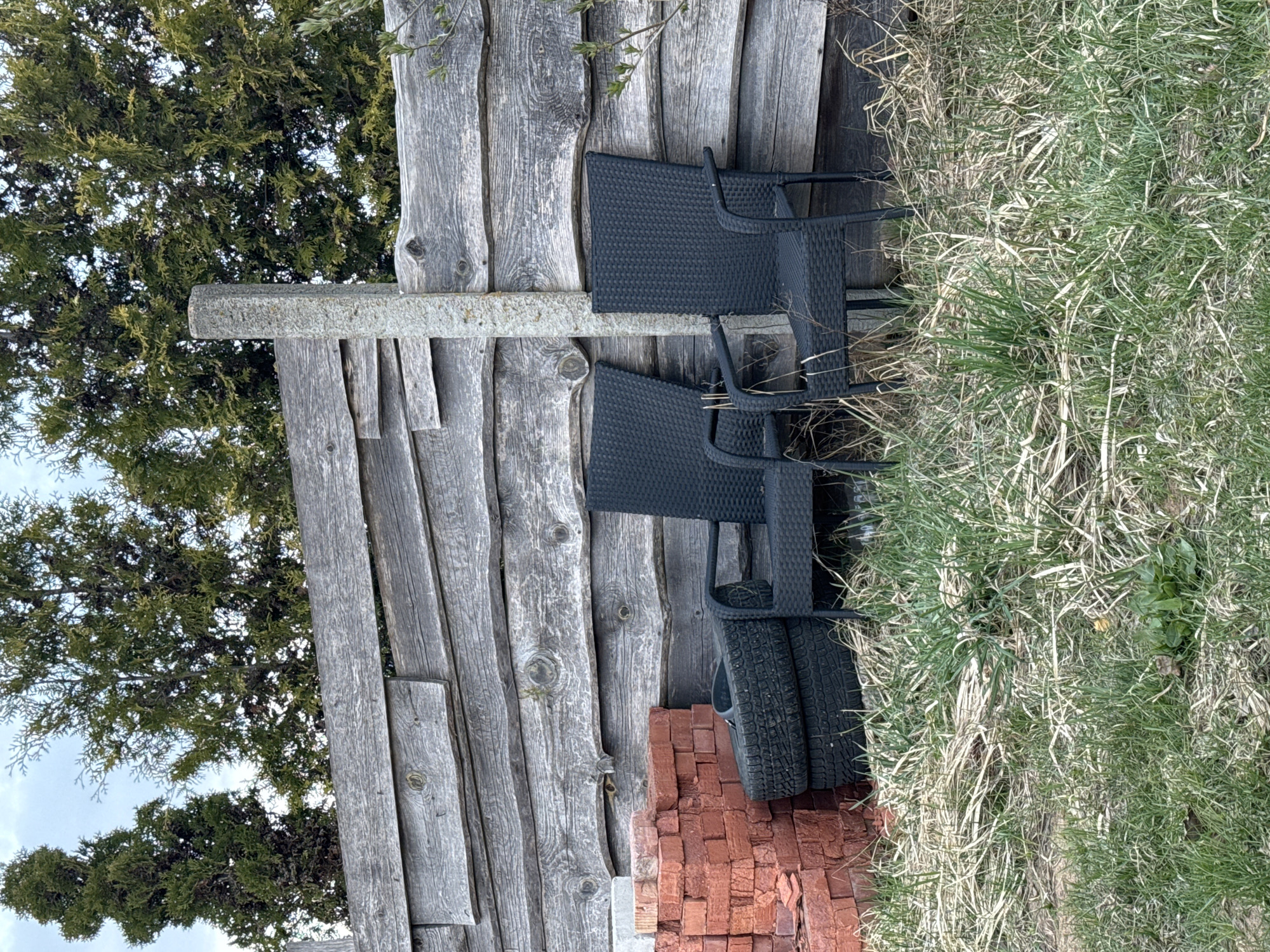 The image displays an outdoor scene in Самахвалавіцкі сельскі Савет, Belarus, featuring a weathered wooden fence with various items stored against it. The fence, constructed from rough, greyed timber planks, stands vertically. A horizontal, rectangular concrete beam is affixed to the fence at mid-height.
Against the fence, two black outdoor chairs, made of a woven material, are stacked. Below them, two black car tires with distinct treads lean against the wooden structure. To the right of the tires, a pile of reddish-brown bricks is neatly stacked on the ground.
The ground area is covered with a mix of green and dry, straw-like grass, suggesting an unkempt patch or an area in transition between seasons. In the background, dense coniferous trees with dark green foliage are visible under a light, possibly overcast, sky. The light indicates it is daytime. No people are present, and no specific activity is taking place. There is no visible text within the image. The scene portrays a utilitarian outdoor space, possibly a backyard or a storage area.