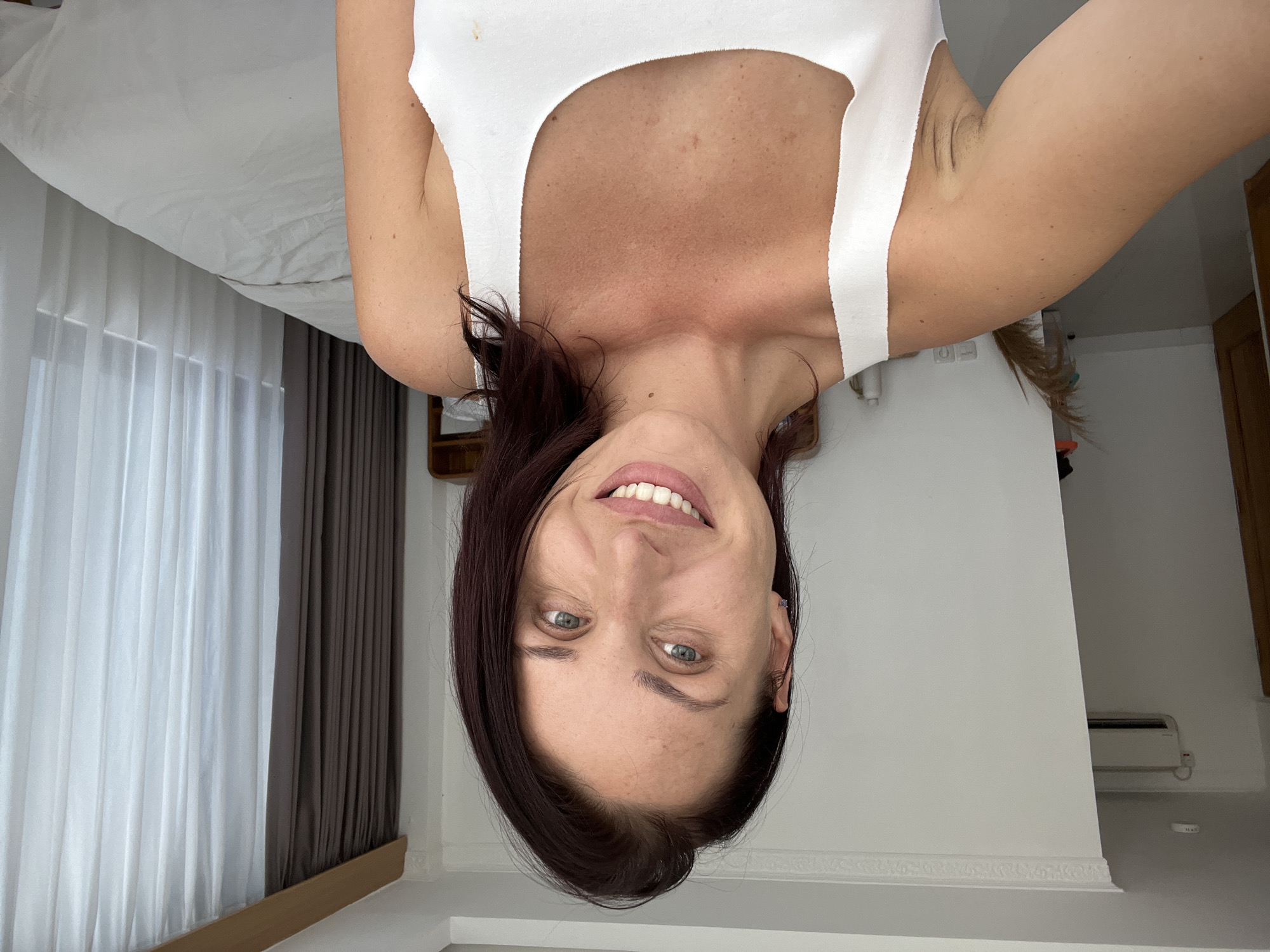 The media file presents an upside-down selfie of a woman in an indoor setting, likely a bedroom, located in Kuta Selatan, Indonesia. The woman is smiling broadly, looking directly at the camera, and has dark hair and light-colored eyes. She is wearing a simple white tank top.
The background of the image, which is also inverted, provides details about the room. To the left, a bed with white bedding is visible, suggesting a minimalist or clean aesthetic. Adjacent to the bed are windows covered by two layers of curtains: a sheer white layer and a darker, opaque grey or brown layer. On the right side of the frame, a plain white wall is visible, featuring a mounted air conditioning unit towards the ceiling and decorative molding along what appears to be a lower wall section.
Notable details include the woman's clear, bright smile. Her skin shows some freckles or sun spots on her chest and shoulders. A cropped view of her raised arm reveals some armpit hair. The overall bright illumination of the room suggests the image was taken during daytime. No visible text is present in the image.