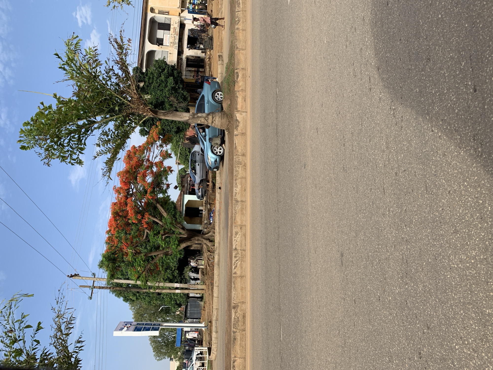 The image shows a street in Rafin Par, Nigeria. Two light blue cars are parked along the curb. A building with multiple levels is in the background with people in front of it. Trees line the street, one having orange blossoms. There are utility poles with wires overhead. The street surface is asphalt. People are walking on the far side of the street. A sign is on the left side of the street.