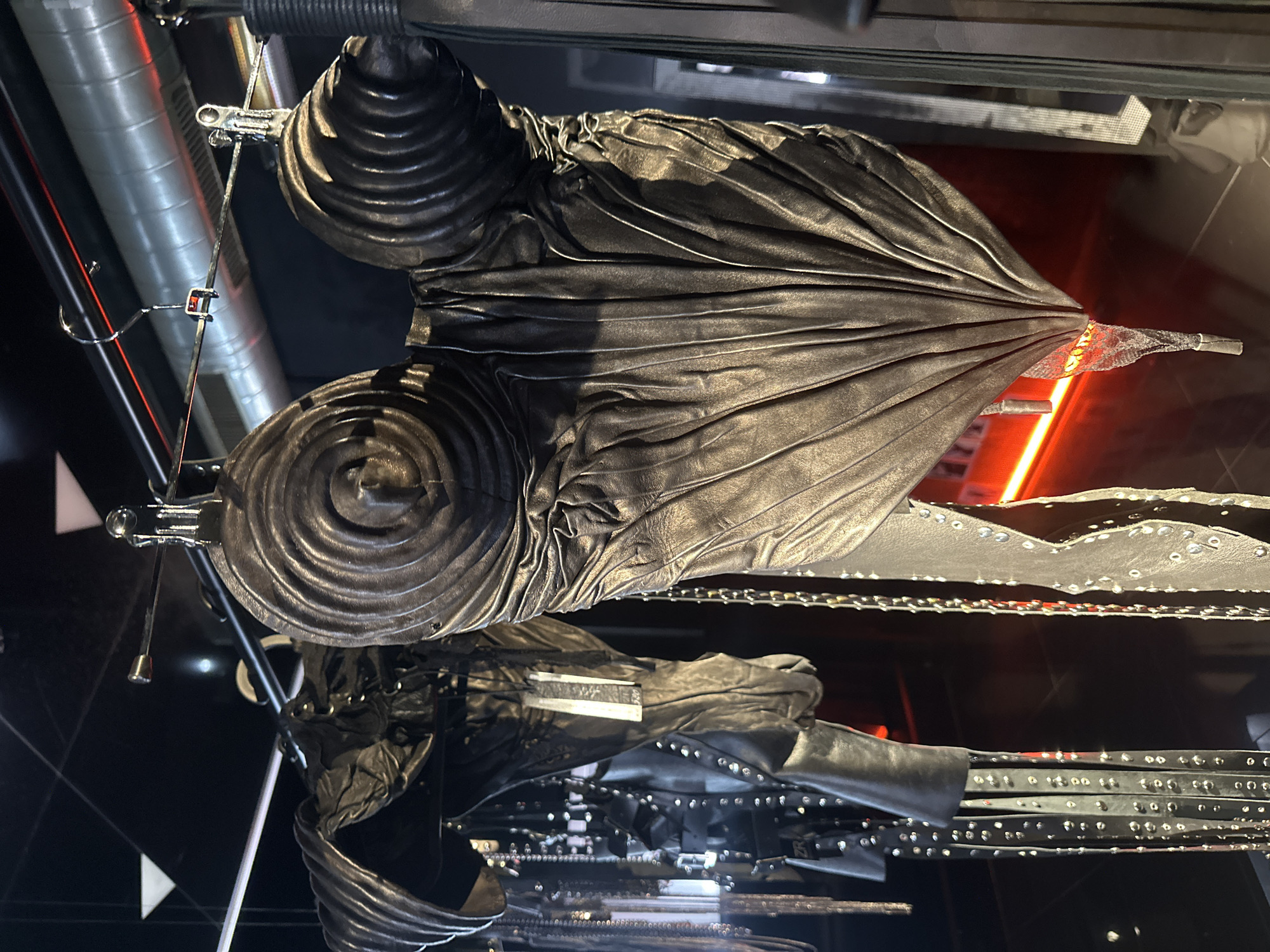 A black leather garment is suspended from a metal rack in an indoor setting. The garment features a textured, swirling design on the upper portion, with a pleated, fan-like section extending outwards. The lower part of the garment has straps or belts adorned with metal studs or rivets. The background includes industrial-looking metal pipes and beams, along with diffused red lighting. The location is Unknown, Unknown.