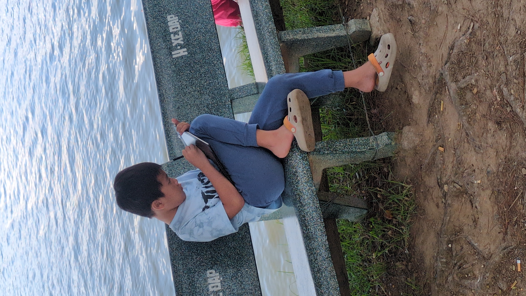 A young person sits on a bench by a body of water, looking at a tablet. The person is wearing a light blue t-shirt and dark blue sweatpants. They have on a beige Croc on their right foot with an orange strap. Their left foot is bare. The bench is made of concrete and has visible text on its side that reads "H-XE-DAP". The water is choppy and appears to be a lake or river. Behind the bench, there is grass and dirt with some exposed tree roots and cigarette butts. The lighting suggests it is daytime, possibly late afternoon given the soft light. The overall scene appears peaceful, with the person engrossed in their tablet, isolated by the water. The location is identified as Da Lat, Vietnam.