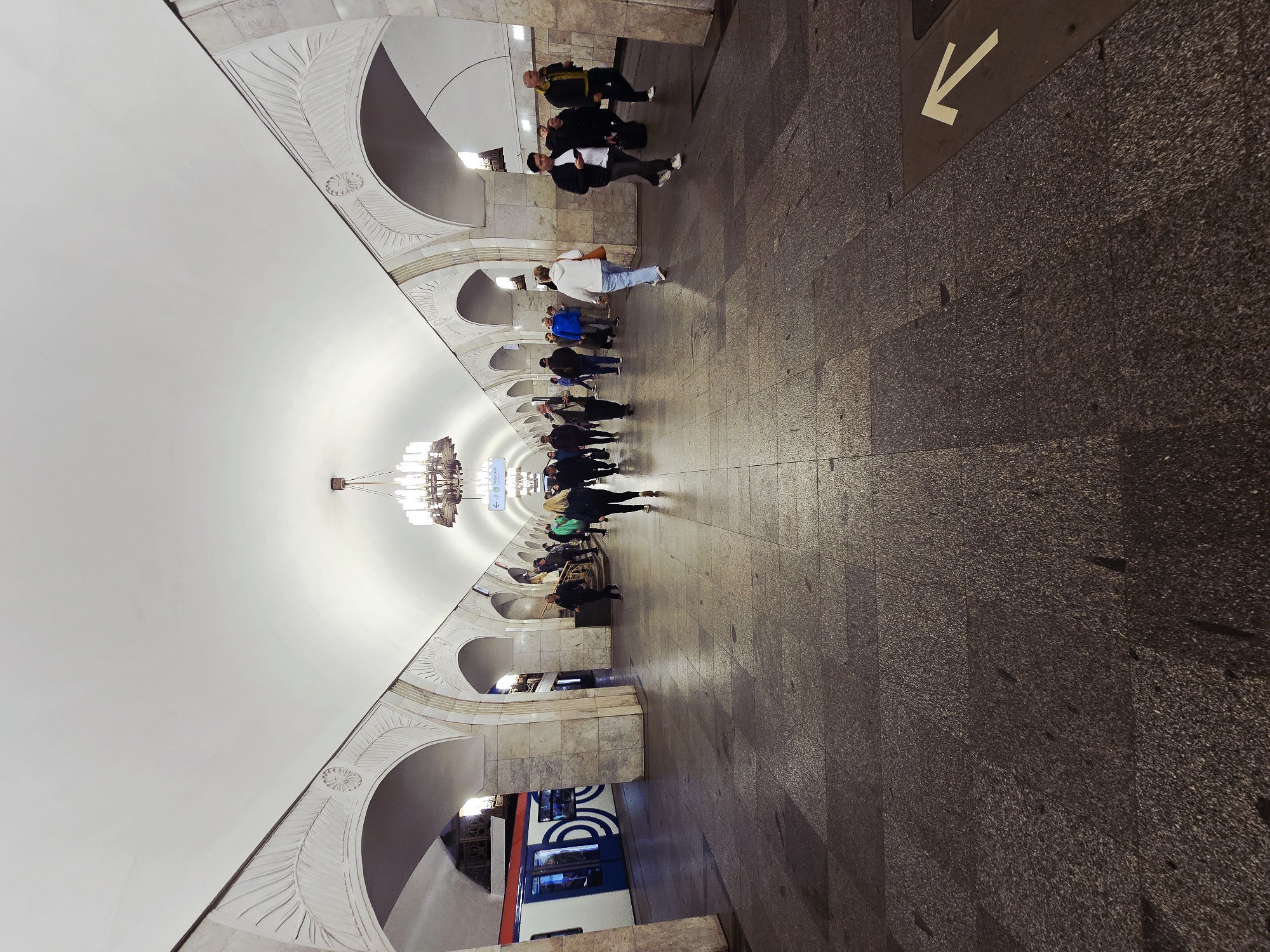 The image depicts a Moscow subway station, most likely the Komsomolskaya station on the Koltsevaya line. The station's architecture is visible with its high, arched ceilings adorned with intricate decorative patterns and grand chandeliers. People are present in the station, some walking along the platform and others waiting for the train. A train is visible on the left side of the image. The floor of the station is made of dark stone tiles. There is an arrow marking the direction of travel on the ground.