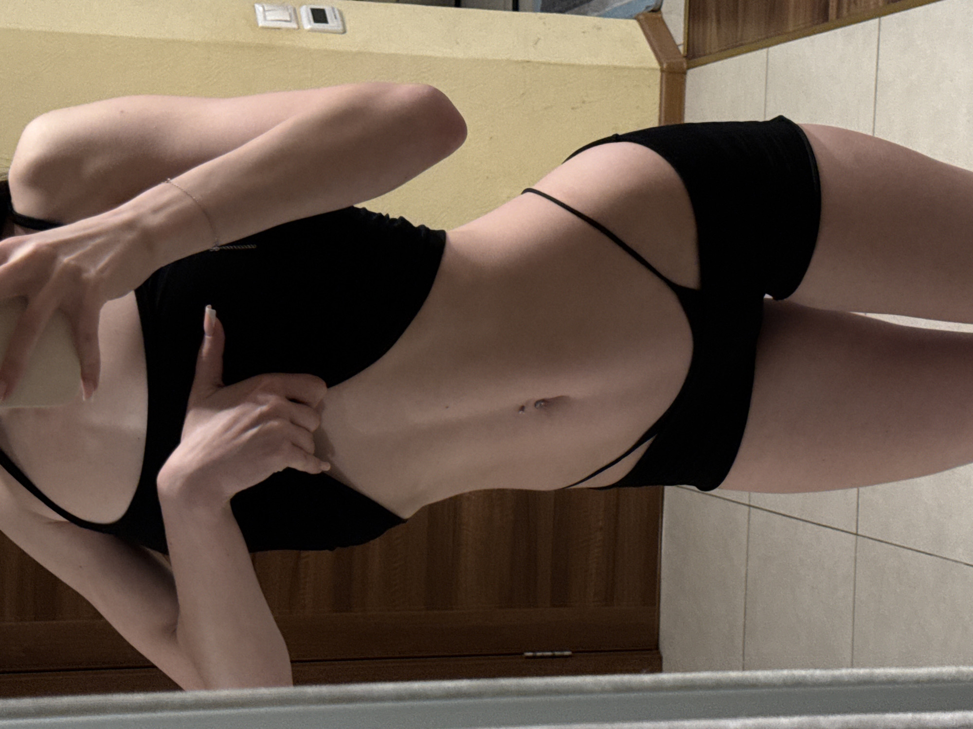 The image shows a close-up, mid-body shot of a person wearing black apparel, in what appears to be an indoor setting. The individual's torso and upper thighs are visible, with a black tank top and a distinctive black bottoms that feature cutouts on the sides. The person's right arm is bent, with their hand reaching under the tank top. A silver bracelet is visible on the right wrist. The environment includes a reflective surface on the left, likely a mirror, showing a sliver of a wooden door and wall. The background wall is a pale yellow, with a light switch visible on the right. The floor appears to be tiled. The image does not contain any identifiable landmarks of the city of Khimki, Russia.