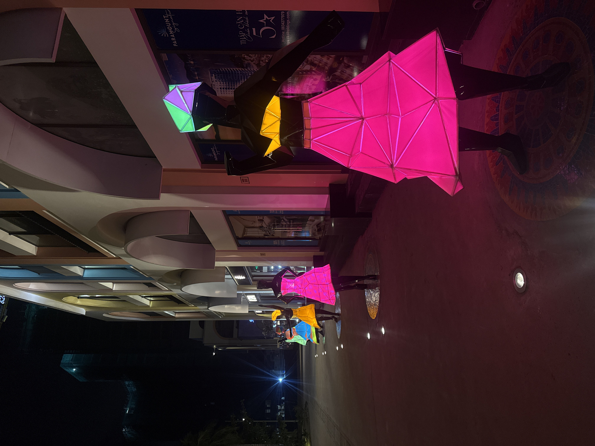 This image depicts a nighttime street scene in the city of Bac Nha Trang Ward, Vietnam, likely a commercial or entertainment district. In the foreground, a mannequin is illuminated by vibrant pink light, wearing a geometric, folded skirt and a polygonal top with green and purple accents. The mannequin's lower body is clad in dark, boot-like coverings, and it stands on a circular, patterned mat. Further down the pathway, a line of similar mannequins are visible, each adorned with brightly colored, geometric dresses in shades of pink, yellow, and blue. The mannequins are spaced out along a paved walkway, which is lit by a series of circular, embedded lights. To the right, a modern building with large arched windows and a light-colored facade is partially visible. A large advertisement poster featuring text and images related to a "Paramount Signature" project is displayed on the building's exterior. The background is dark, with the silhouette of a tall, under-construction building visible against the night sky. The overall atmosphere is artificial and vibrant, illuminated by the colorful lights of the mannequins and the building's lighting.