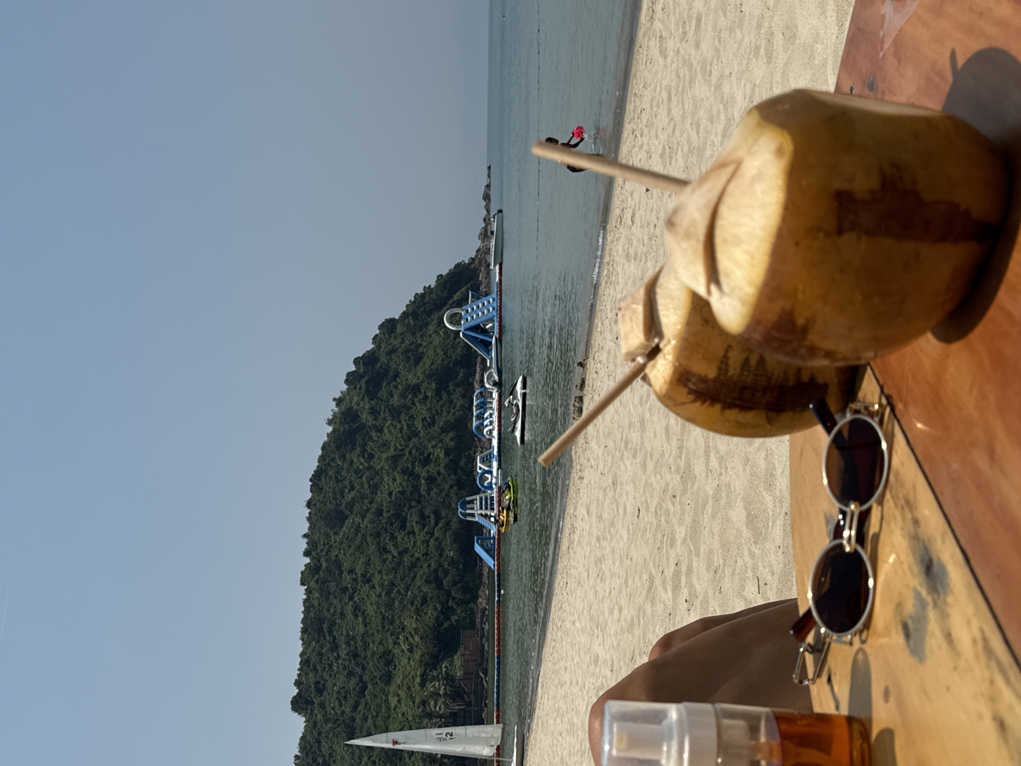 The image depicts a first-person perspective from a shaded spot on a beach, looking towards the ocean and a forested mountain. In the immediate foreground, a person's bare legs are visible on the left. On a light-brown wooden surface, two light-orange coconuts with straws stand upright; one has a dark vertical stain on its side. Between the coconuts and the legs, a pair of dark-lensed, circular sunglasses with a silver-colored frame rests on the table, alongside a clear plastic spray bottle with orange liquid and a white foam top.
Beyond the table, a wide expanse of light-colored sand leads to the calm, greenish-blue ocean. In the water, a large blue and white inflatable water park with slides and climbing structures is prominent. Several watercraft are present: a white sailboat with sail number "112" near the left shore, two personal watercraft (jet skis), and a small white motorboat further out. A line of buoys separates areas of the water. On the right, a dark-skinned individual wearing dark clothing wades in the shallow water near the shore, holding a pink object.
The background is dominated by a lush, dark green, tree-covered mountain or hill that extends down to the water's edge on the left. The sky above is clear and light blue. The scene is brightly lit by natural sunlight, casting shadows. This location is a beach situated on the Sơn Trà Peninsula in Đà Nẵng, Vietnam.