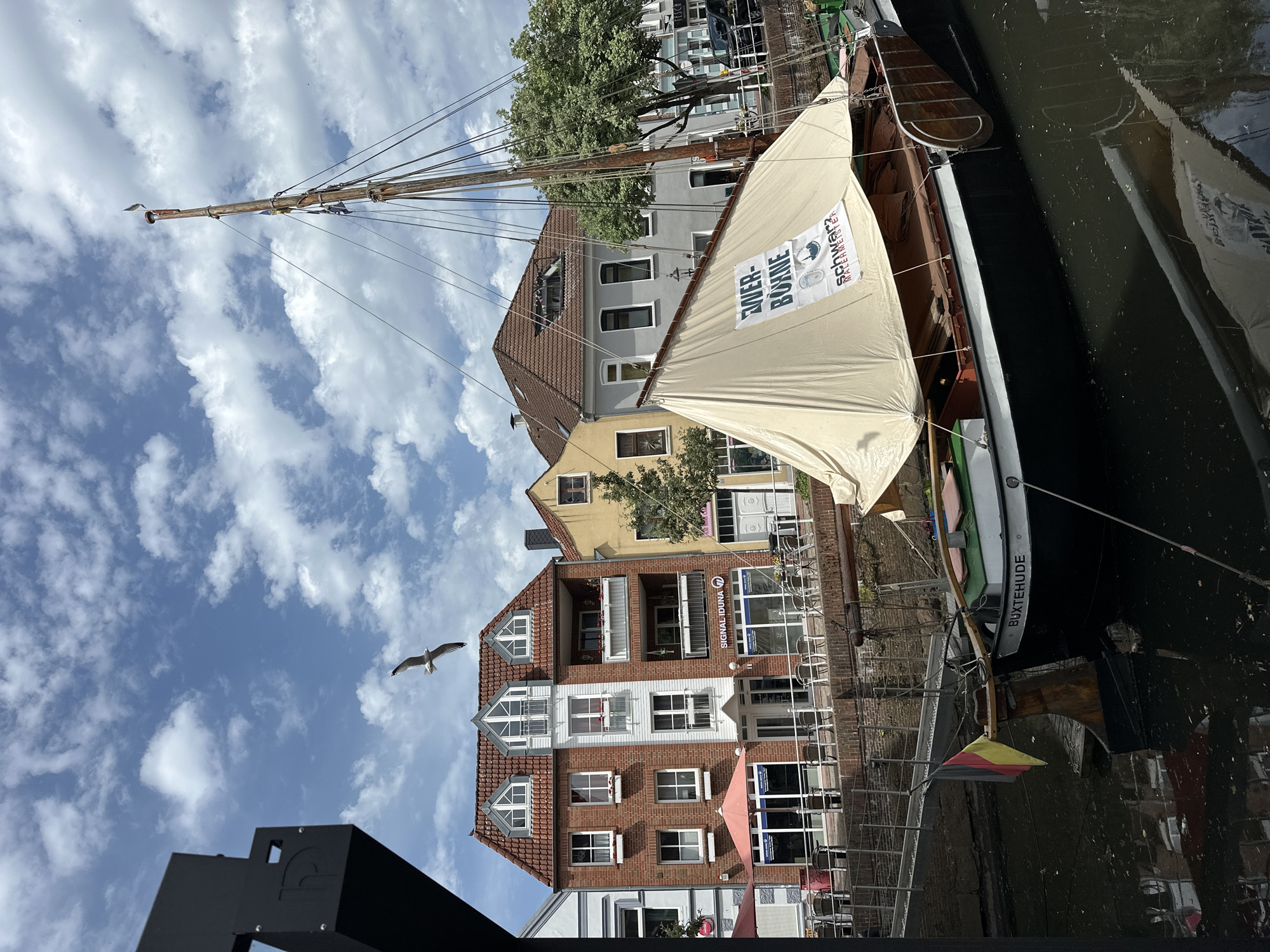 A view of a body of water, possibly a canal, with a historical sailing boat docked alongside. The boat has a light tan sail with "WER-BUMME" and an image on it. The stern of the boat bears the name "BUXTEHUDE", identifying the location as Buxtehude, Germany. Buildings stand in the background, with red brick and tan facades. A bird flies across the sky. The sky is blue with scattered clouds. Parts of a black metal structure are visible in the bottom left corner of the frame.