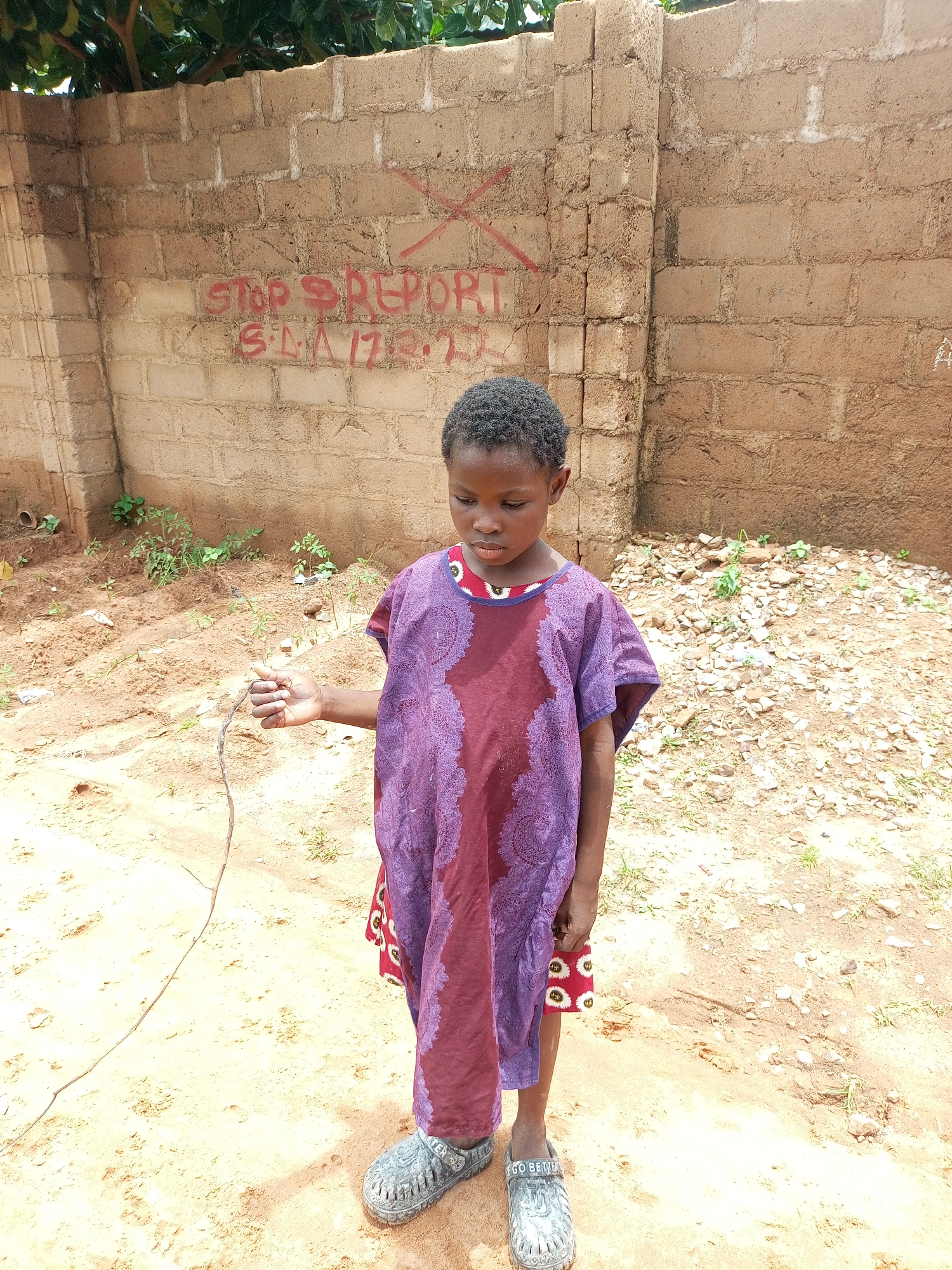 A young person stands in front of a rough, tan brick wall in what appears to be an outdoor area in Tempure, Nigeria. The wall has red graffiti on it, including what looks like an "X" and the words "STOP REPORT" and "S.L.A. 17.8.21." The person is wearing a purple and maroon patterned dress and gray, perforated shoes. They are holding a thin, dry stick in their right hand. The ground is sandy with some scattered rocks and small green plants. Trees with green leaves are visible at the top of the image. The lighting suggests it is daytime.