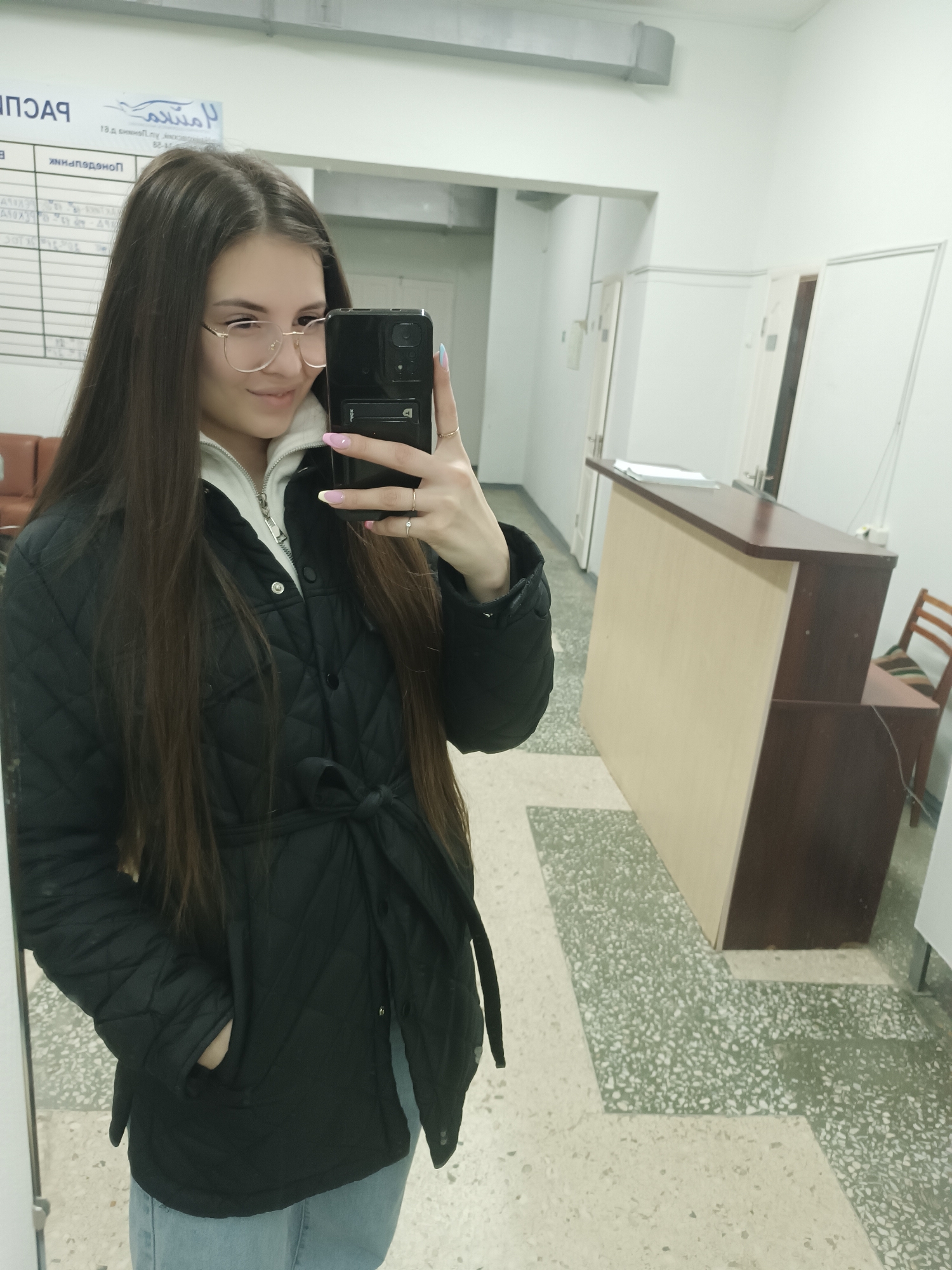A young woman is taking a selfie in what appears to be an office or medical facility. She has long, dark hair, glasses, and is wearing a black, quilted jacket over a white hooded sweater, and light blue jeans. She is holding a black smartphone in her right hand.
The background includes a reception desk, white walls, and a linoleum floor with a speckled pattern. There is a notice board or chart on the wall behind her to her left. A chair is visible to the right of the reception desk. The city and exact location are indeterminable from the given image.
