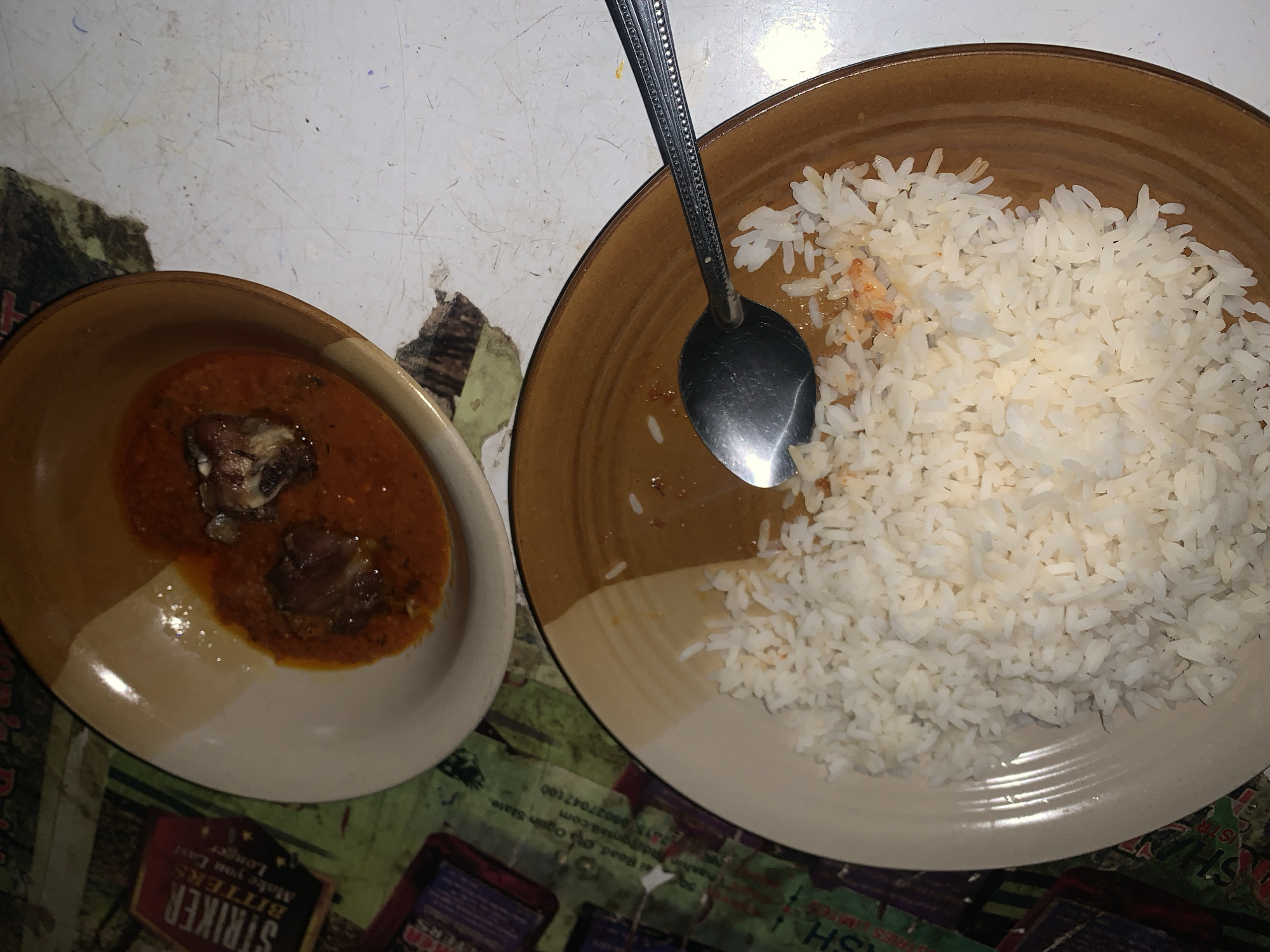 The image shows two plates of food, one containing white rice and a spoon, and the other containing a stew-like dish with pieces of meat. The food appears to be placed on a table with a damaged and stained surface. A portion of a sign is visible beneath the plate with the stew, mentioning "Striker Bitters" and the address "41 Road Ota Ogun State" indicating a location near New Stadium, Nigeria, based on the address information.