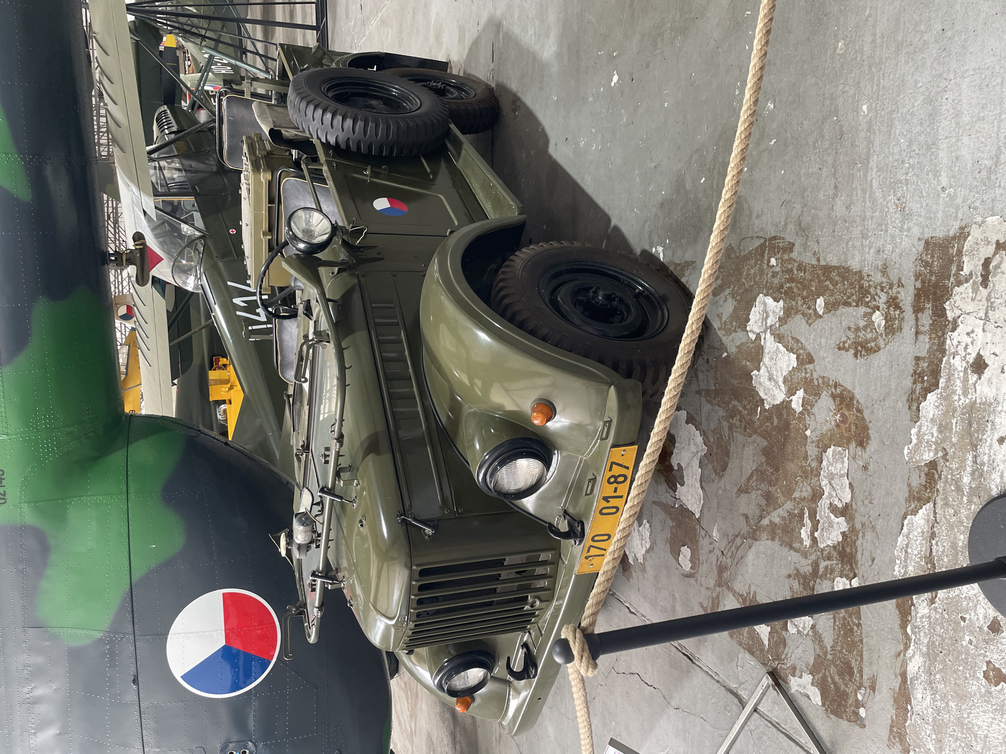 An olive-green, open-top military jeep is parked on a concrete floor in what appears to be a museum or exhibition hall. The jeep is positioned at an angle, with its front facing slightly to the left. It has a spare tire mounted on the right rear side, and a Czech flag emblem is visible on the right fender. A yellow license plate with the number "170 01-87" is affixed to the front bumper. Behind the jeep, a large aircraft, also painted in camouflage, is partially visible. The aircraft has a circular emblem on its side, which is also red, white, and blue. A rope barrier is set up in front of the jeep, and a dark-colored pole stands to its left. The floor shows signs of wear and tear, with patches of peeling paint or concrete. The lighting suggests an indoor environment, possibly with overhead lights.