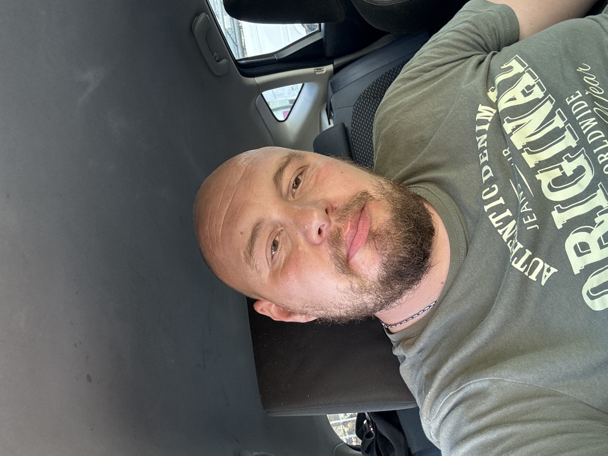 A man with a shaved head and a beard is taking a selfie inside a vehicle. He is wearing a green t-shirt with a design on it. The interior of the car is visible, including the seats and the roof lining. The image appears to be taken during the daytime. There is no visible indication of the location being specifically within the city of Cork, Ireland.