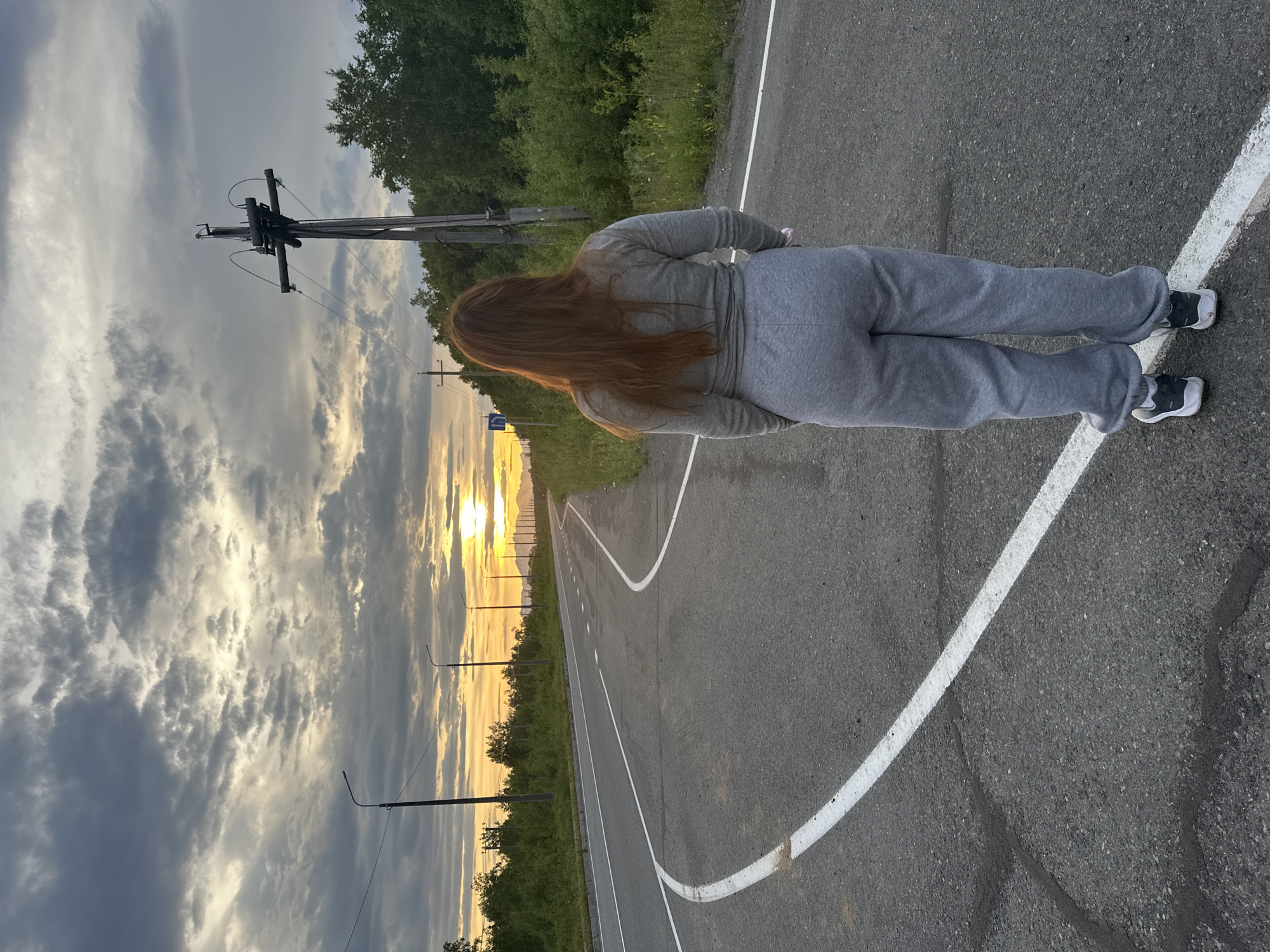 A person with long, red hair stands on a paved road at dusk, facing away from the camera. They are wearing a grey sweatshirt and grey sweatpants, with black and white sneakers. The road curves to the left and continues into the distance. On the right side of the road, there is a wooden utility pole with wires. Dense green trees line the right side of the road. The sky is filled with dramatic, grey and yellow clouds, with the sun setting on the horizon, casting a warm glow. The location is identified as Ketkino, Russia.