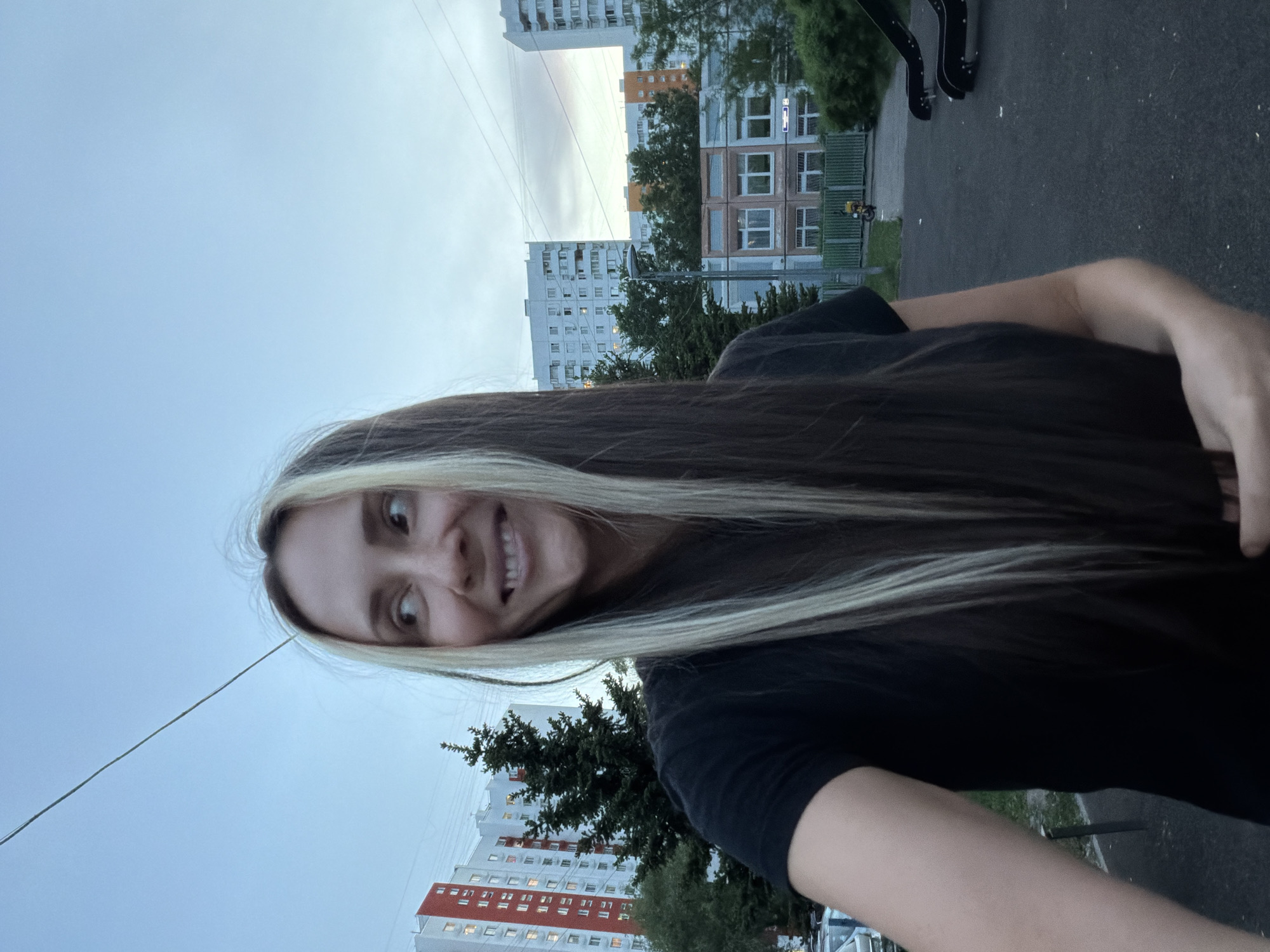 A young woman takes a selfie in what appears to be a residential area in Moscow, Russia. She is smiling and has long, dark hair with blonde streaks. In the background are apartment buildings typical of the area, a green space with trees, and a cloudy sky. There is also a power line visible. The photo seems to be taken outdoors during daylight, possibly in the evening based on the sky's color.