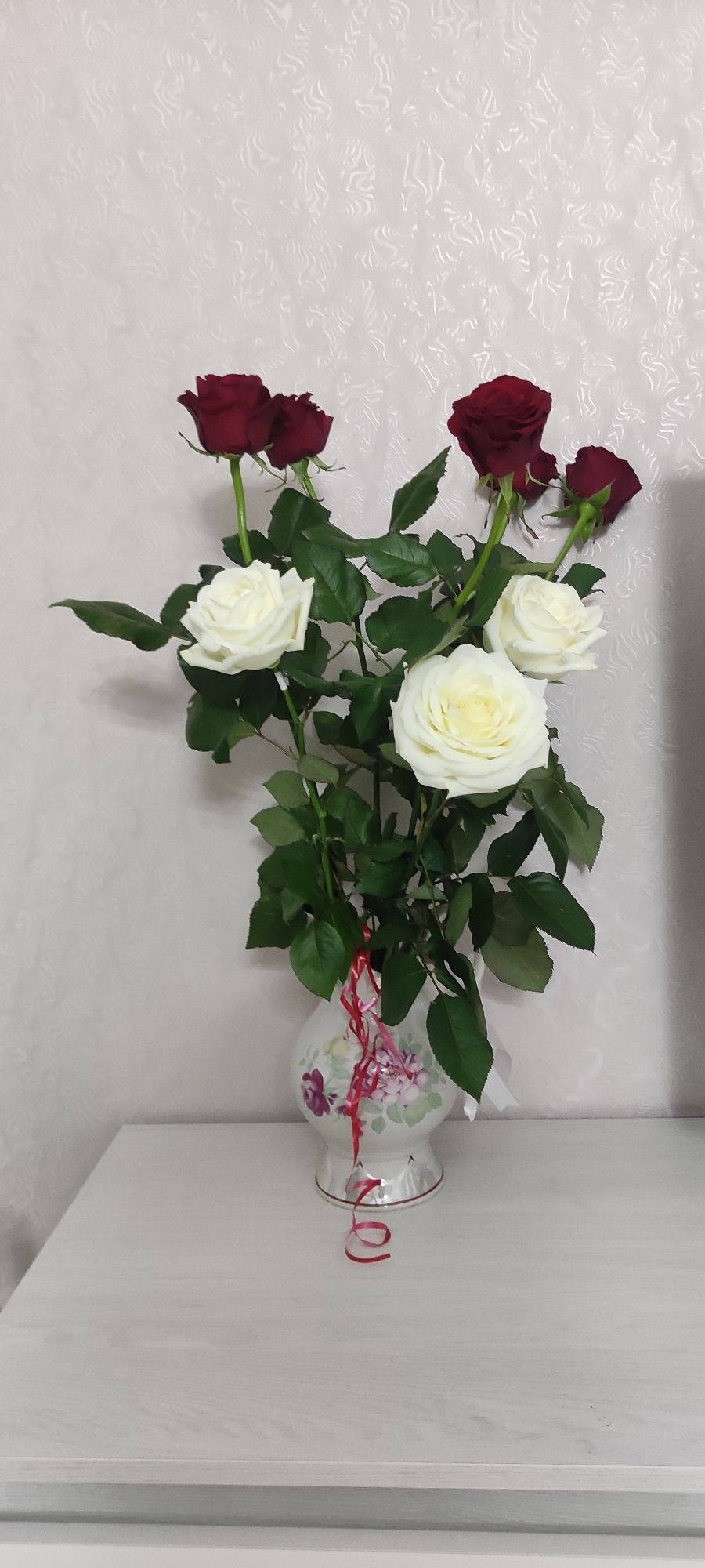 A vase of red and white roses sits on a light-colored table against a neutral wall. The vase is decorated with a floral pattern. Red ribbons hang from the vase. The location is Chaikovsky, Russia.