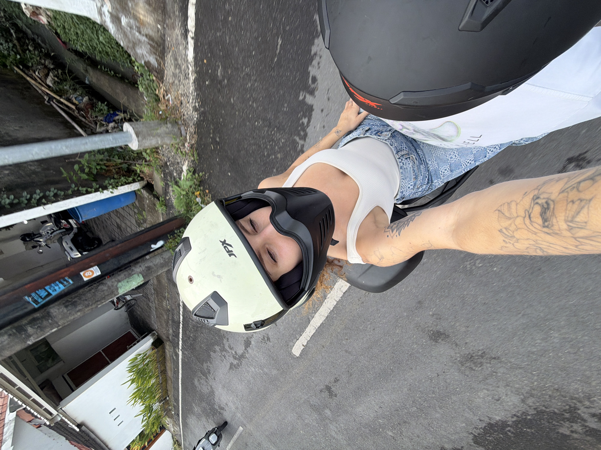 The image shows two people on a motorcycle in North Kuta, Indonesia. The person in the foreground is wearing a light yellow helmet with a black face mask and a white tank top. They are looking down and to the right, with their eyes closed. The person in the background is wearing a black helmet. The motorcycle is on a wet asphalt road, with a white line painted on it. To the left of the road, there is a white building with a red door and some plants. To the right of the road, there is a concrete wall with some greenery and debris. There are also a few other motorcycles visible in the background. The sky is overcast.