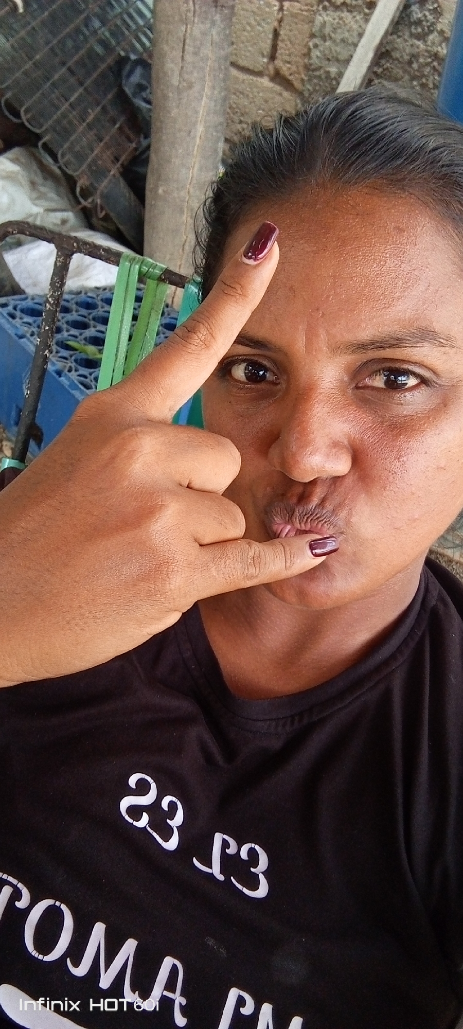 A person with brown skin and dark hair is shown in a selfie style image. They are wearing a black t-shirt with white lettering that reads "23-23 TOMA CAM." The subject is pouting their lips and making a gesture with their hand, extending the index finger toward their lips and their thumb toward their forehead. Both the index finger and thumb have dark red nail polish. The background includes a metal structure, a wooden post, and a blue container. The bottom left corner of the image contains the text "Infinix HOT 6i." The location is Altagracia, Venezuela.