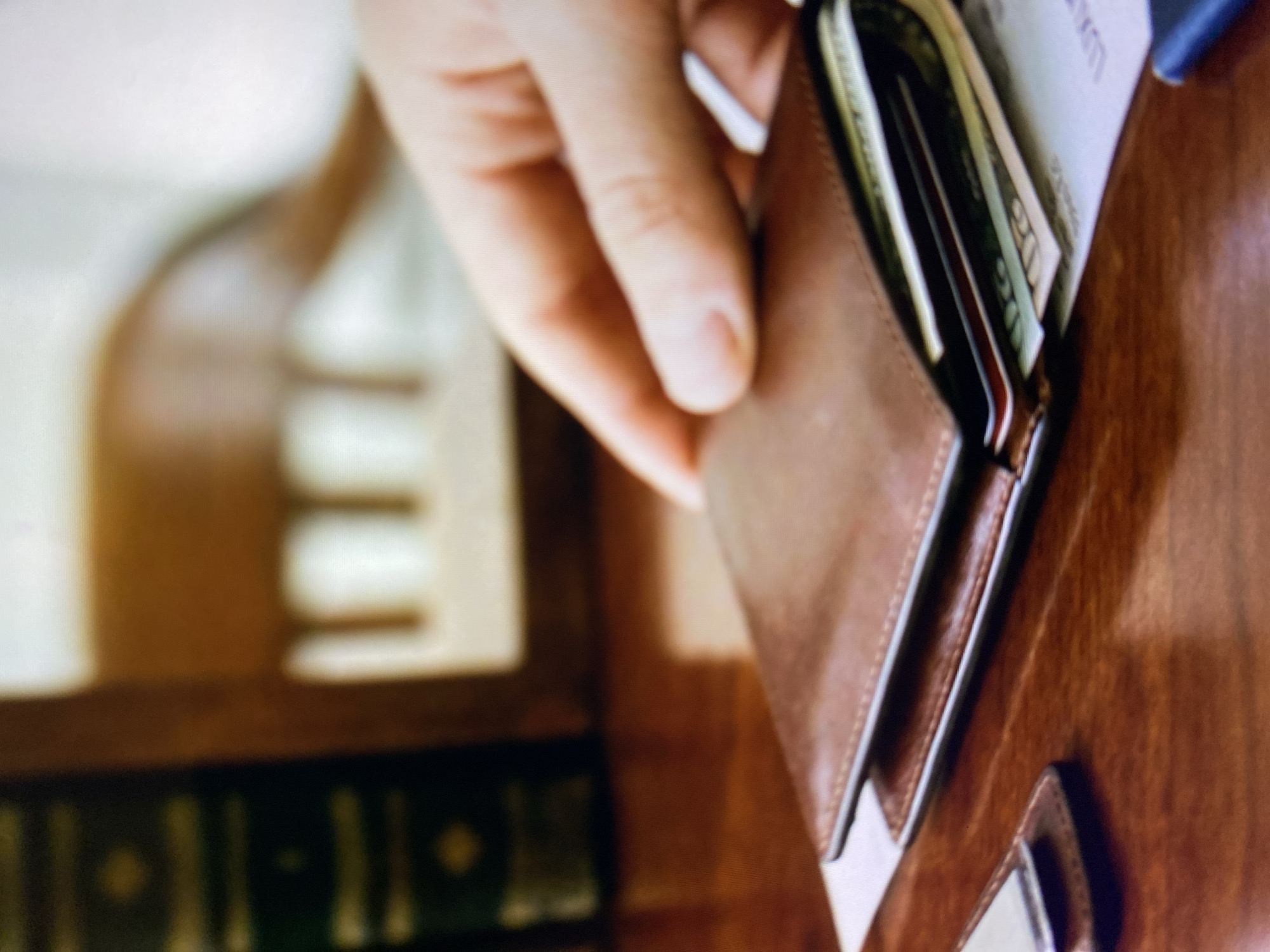 A hand is reaching for a brown leather wallet filled with cash and cards on a wooden desk. The wallet is slightly open, revealing several bills, including what appears to be a $20 bill. A portion of a dark phone is visible on the left side of the frame, also resting on the desk. In the background, out of focus, are books and a chair. There is no information available about the location in the city of Kuta Selatan, Indonesia.