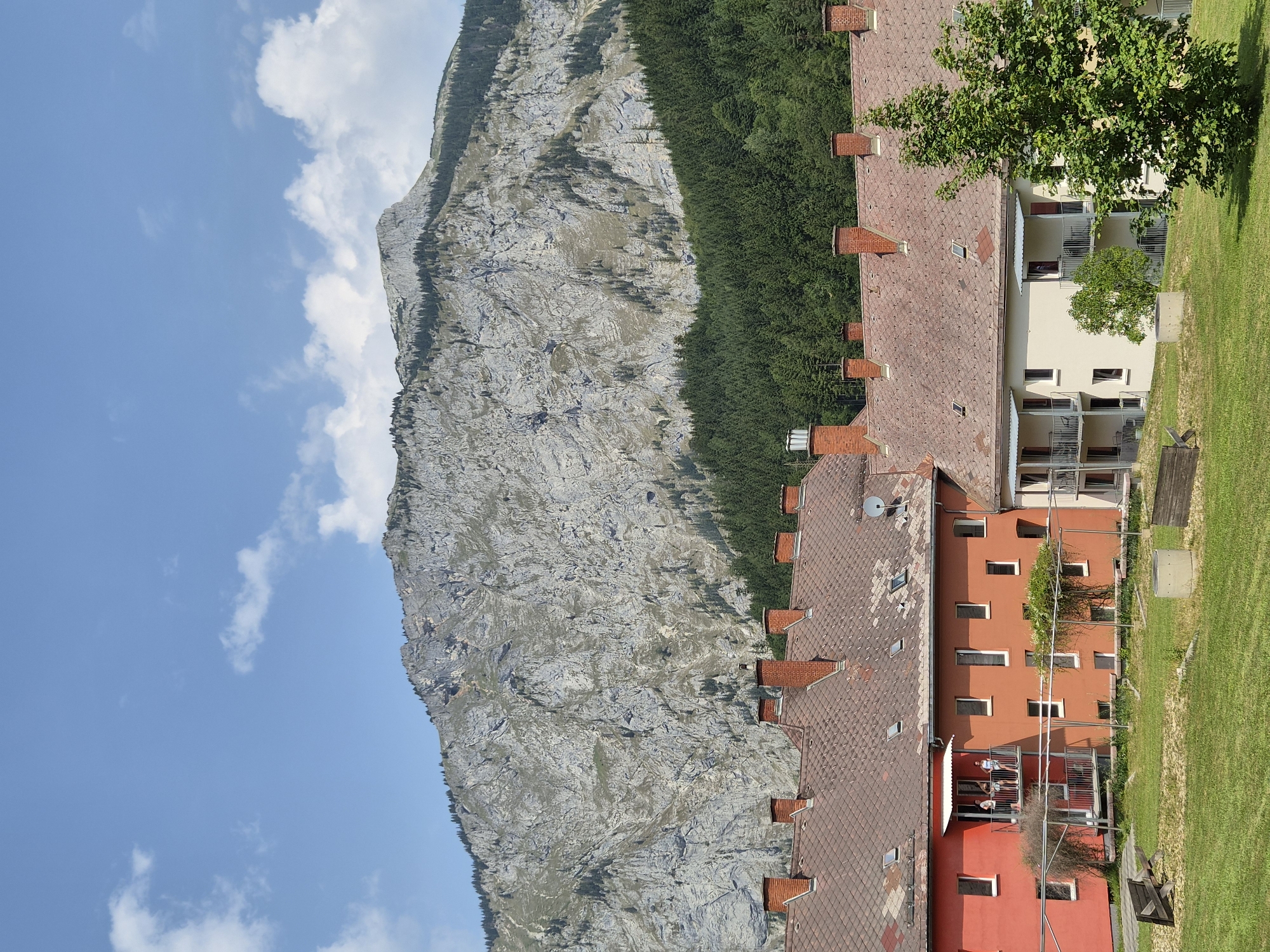 The image depicts a view of buildings with red-tiled roofs, a large rocky mountain face in the background, and a dense forest of green trees situated between the buildings and the mountain. The sky is blue with scattered white clouds. In the foreground, there is a grassy area with a wooden bench and a concrete structure. The buildings appear to be apartment complexes or similar multi-story residential structures. One building on the left is painted bright red, while others are a muted orange and cream color. Balconies are visible on the buildings, and a few people can be seen standing on a balcony of the red building. The overall setting suggests a mountainous region with human habitation, characteristic of towns like Eisenerz, Austria, known for its iron ore mining and surrounding natural landscapes.