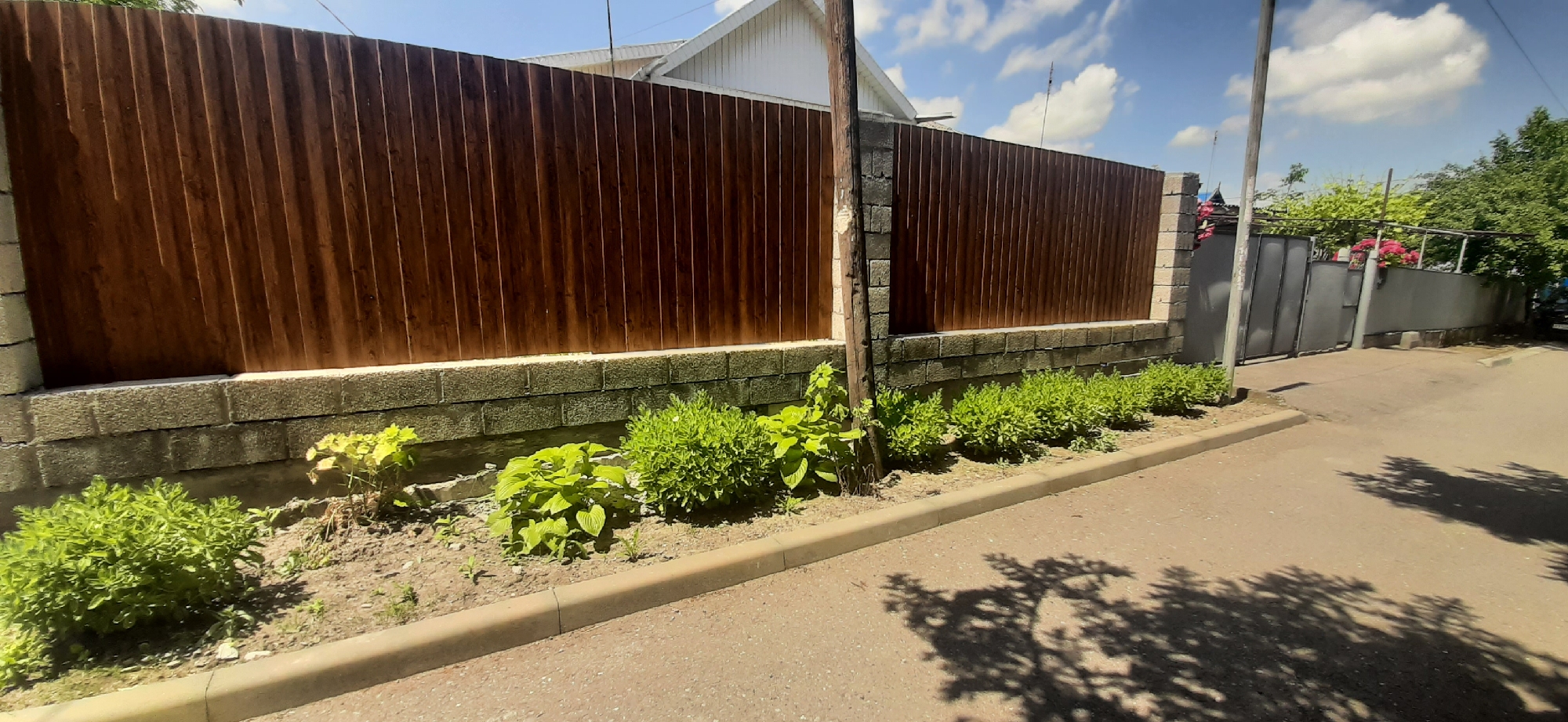 The image shows a street scene in Yessentukskaya, Russia. On the left, a dark brown wooden fence with vertical planks stands atop a foundation of grey concrete blocks. In front of the fence, a line of lush green bushes grows along a sandy strip. To the right of the fence, a grey metal gate is visible, set into a grey concrete wall. Beyond the gate, pink flowers are in bloom, and trees with green foliage are visible. The street is paved with asphalt and a light-colored curb runs along the edge. Shadows of tree branches are cast across the street. The sky is blue with scattered white clouds.